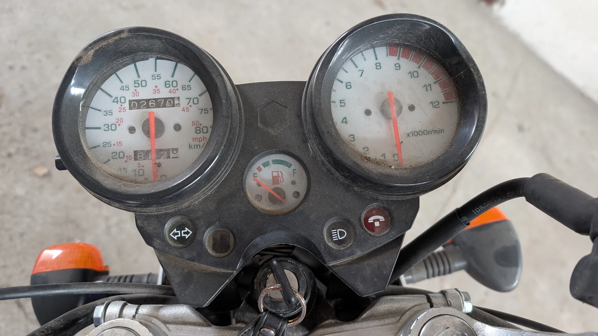 Zbliżenie na deski rozdzielcze motocykla z dwoma głównymi wskaźnikami. Po lewej stronie znajduje się prędkościomierz z zakresem 0-80 mph (dodatkowo oznaczenia km/h), cyfrowy licznik przebiegu pokazujący 02670 oraz czerwona igła wskazująca prędkość. Po prawej stronie tachometr z zakresem 0-12 (oznaczenie x1000r/min) i czerwoną igłą. W centrum mały wskaźnik paliwa z oznaczeniami E (pusty) i F (pełny) oraz ikoną zbiornika. Poniżej wskaźników przyciski: kierunkowskazy (strzałki), światła (symbol D) i inny przycisk z czerwoną ikoną. Widoczne są również pomarańczowe kierunkowskazy na kierownicy, klucz w stacyjce oraz zardzewiałe elementy metalowe. Tło stanowi betonowa powierzchnia. Deska rozdzielcza jest zapylona.
