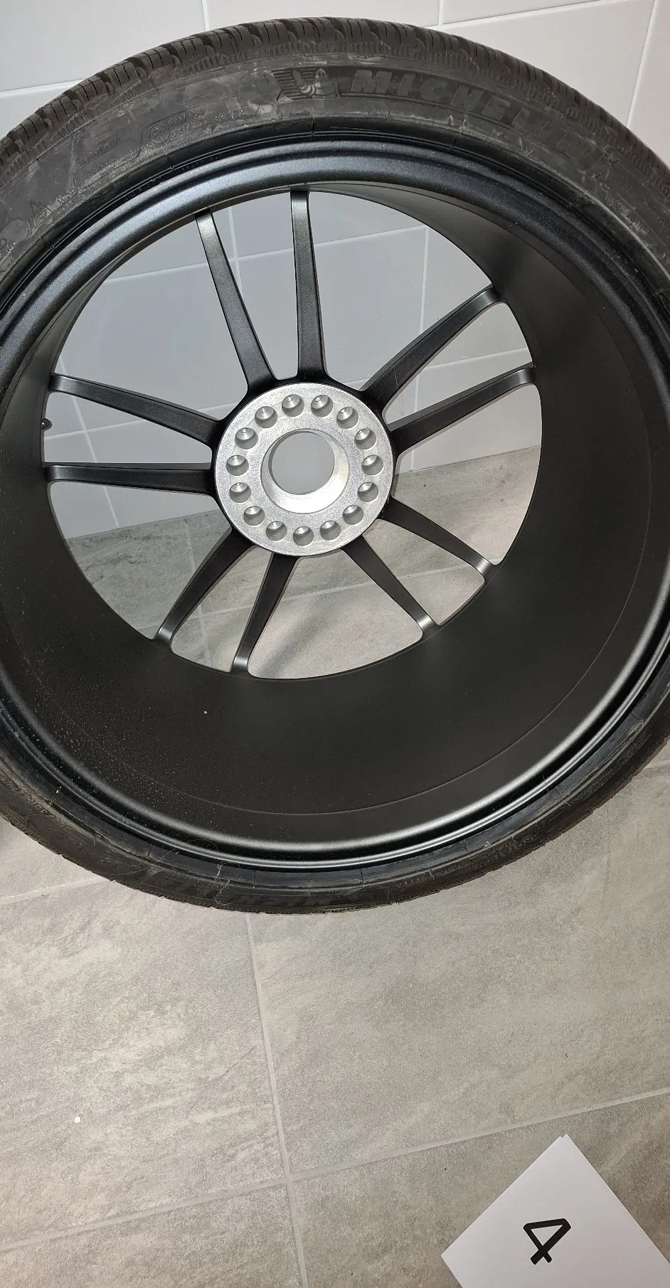 Czarna, matowa felga samochodowa z oponą marki Michelin, oparta o białą ścianę i stojąca na szarych płytkach ceramicznych. Felga ma wiele cienkich ramion rozchodzących się od centralnego pierścienia z otworami na śruby. W prawym dolnym rogu zdjęcia widoczna jest biała kartka z napisem '4'.