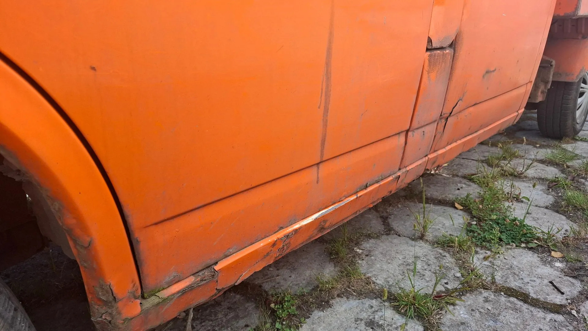 Zbliżenie na dolną część pomarańczowego nadwozia samochodu. Widoczne są liczne uszkodzenia lakieru, wgniecenia oraz zaawansowana korozja z odpryskami rdzy na progu i nadkolu. Pojazd stoi na betonowym podłożu z wyrastającą spomiędzy płyt trawą.