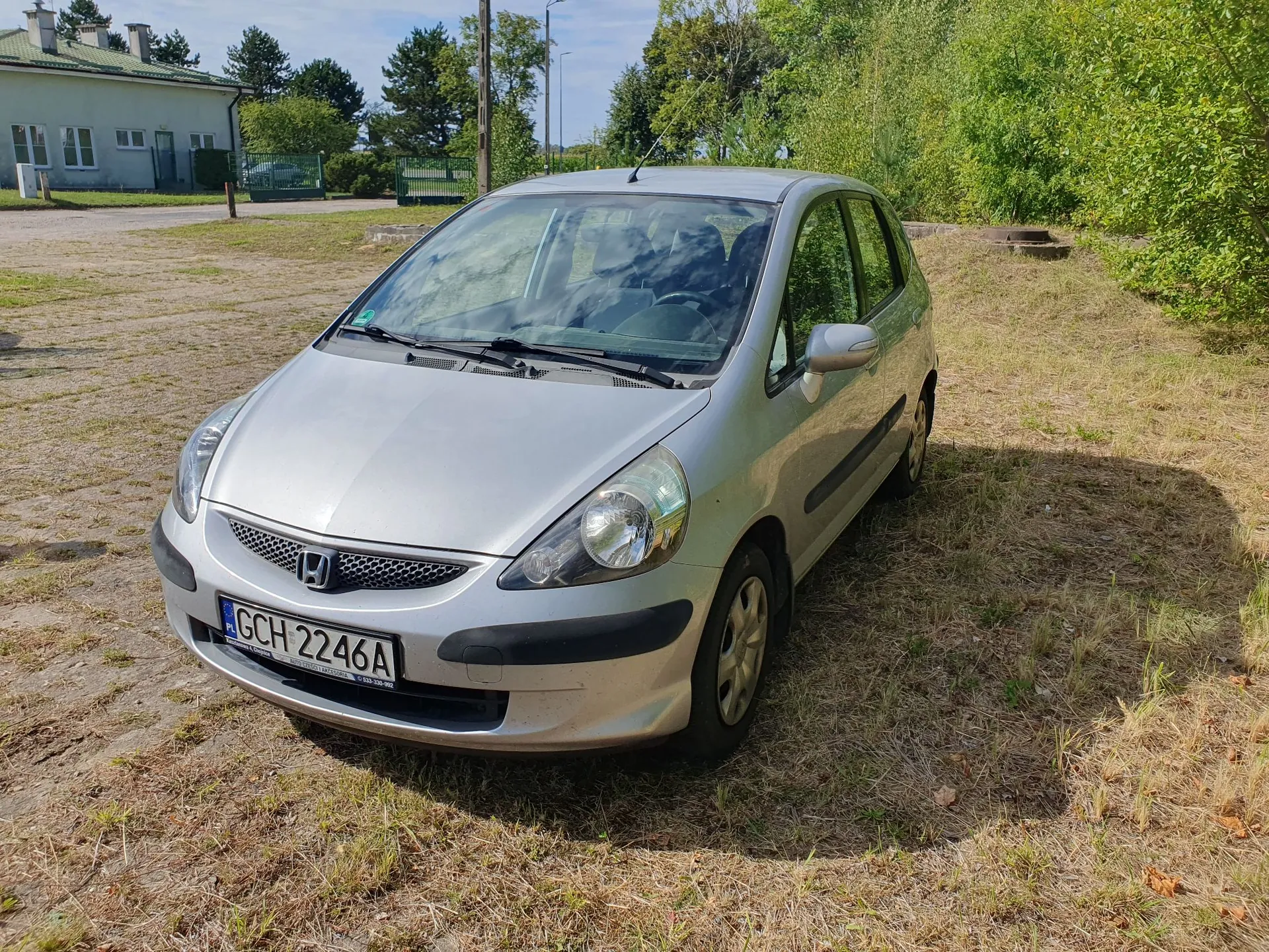 Srebrny samochód osobowy marki Honda zaparkowany na trawiastym i piaszczystym terenie. Pojazd ma polską tablicę rejestracyjną o numerze GCH 2246A. W tle widoczny jest biały budynek z zielonymi drzwiami oraz drzewa i krzewy pod jasnym, niebieskim niebem.