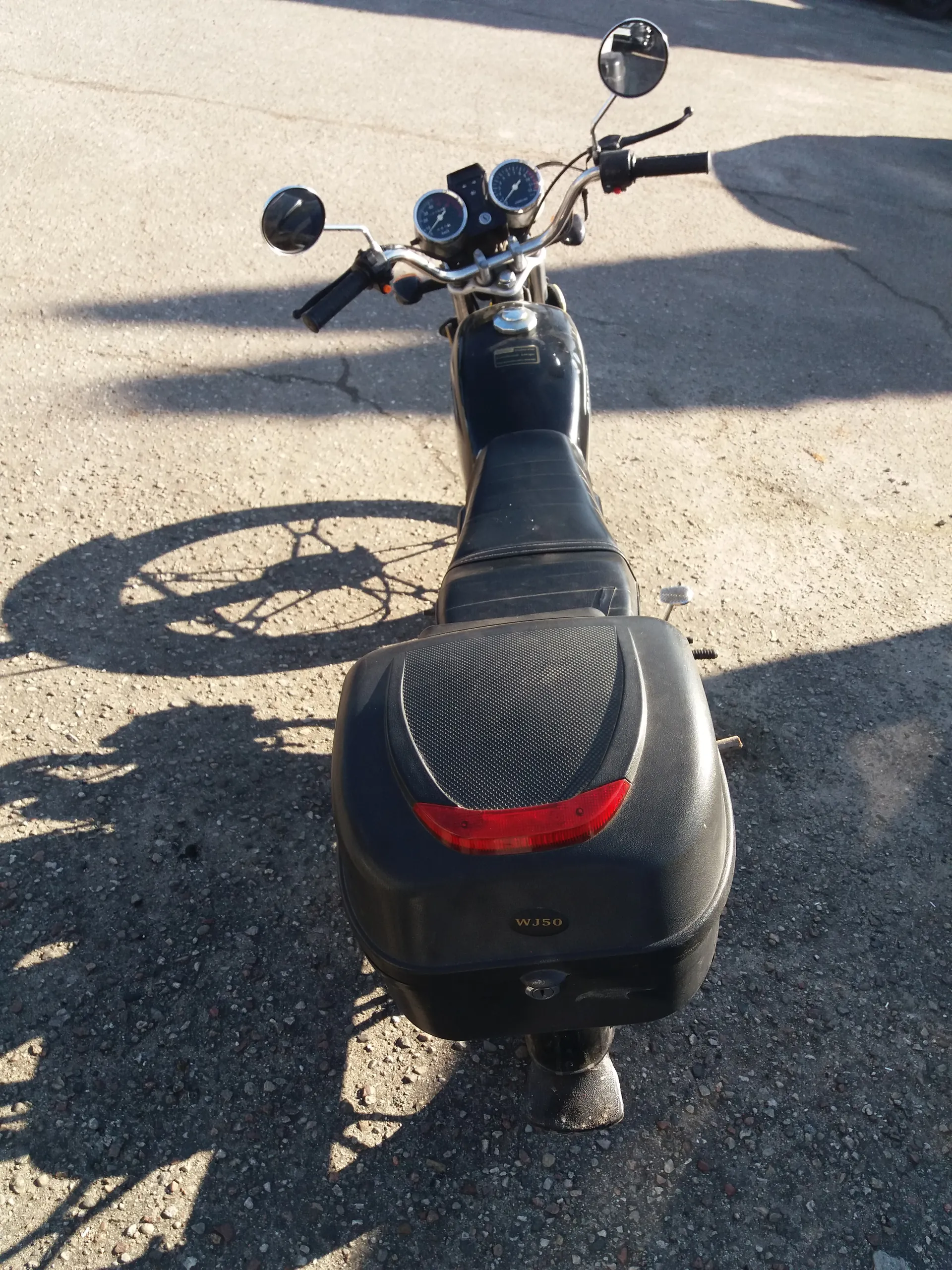 Czarny skuter widziany z góry, stojący na asfaltowej powierzchni. Skuter posiada czarną siedzisko, kierownicę z dwoma lusterkami, wskaźnikami prędkości oraz czarną skrzynię bagażową z czerwonym światłem tylnym. Na skrzyni bagażowej widoczny jest napis 'WJ50'. W tle asfalt z widocznymi cieniami.