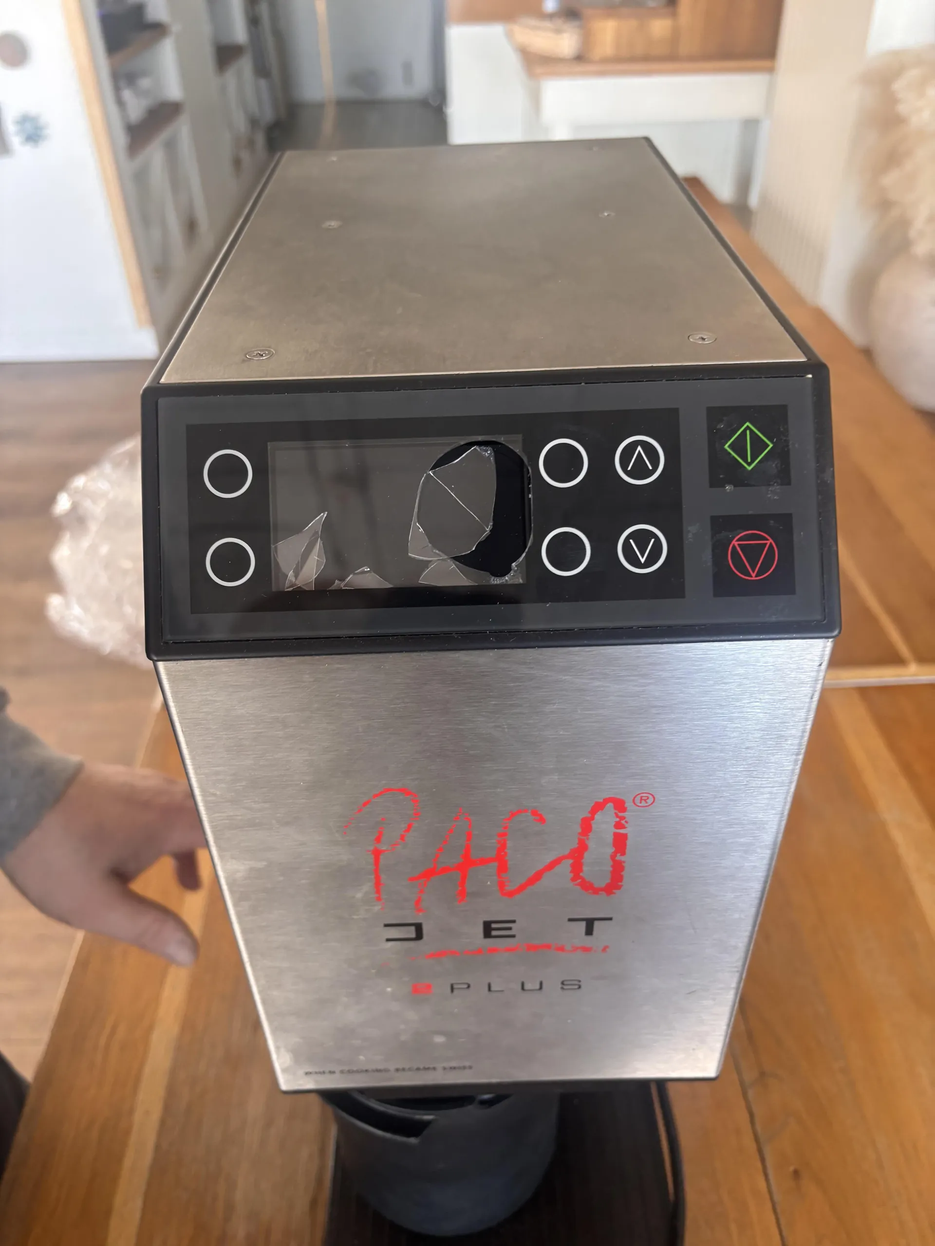 Stalowa maszyna z czarnym panelem sterowania, na którym widoczne są przyciski: cztery okręgi, strzałki w górę i w dół, zielony romb oraz czerwony trójkąt. Ekran na panelu jest pęknięty. Na przedniej części maszyny czerwonymi literami napisane 'PACO JET' oraz poniżej 'PLUS'. W tle drewniana podłoga, fragment ręki trzymającej maszynę oraz meble kuchenne. Obok maszyny widoczny jest przezroczysty worek z plastiku.