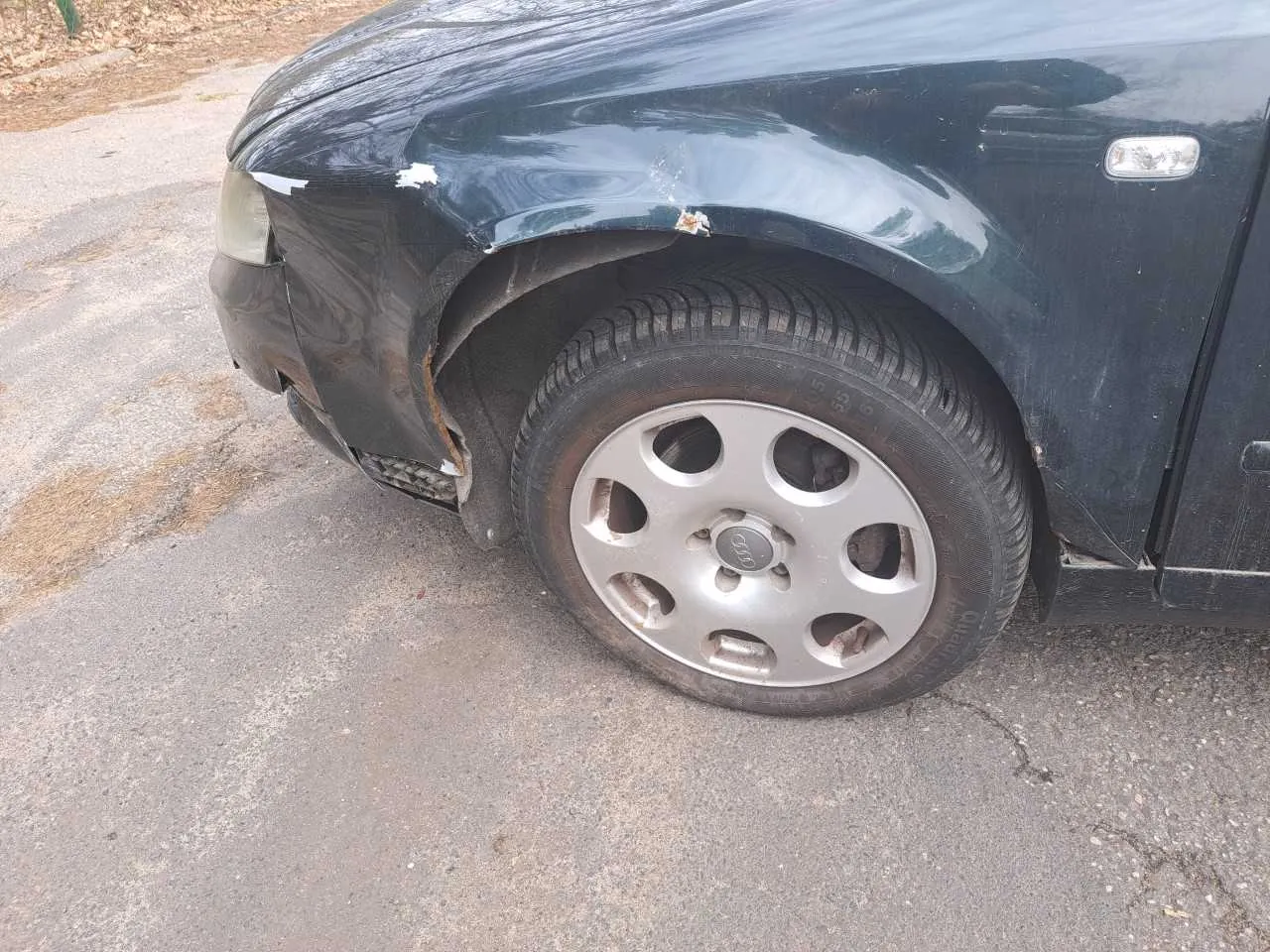 Czarny samochód marki Audi z widocznym uszkodzeniem przedniego lewego zderzaka i nadkola. Widoczne są srebrne felgi z logo Audi, opona z napisem 'Continental' oraz asfaltowa powierzchnia z pęknięciami. W tle fragment suchych liści i brzegu chodnika.