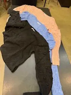 Cztery swetry ułożone na szarej powierzchni: czarny z guzikami, niebieski, jasnoniebieski i brzoskwiniowy. W tle widoczna część podłogi z drewna. Brak czytelnego tekstu.