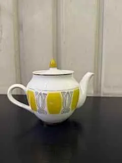 Biały czajnik z żółtymi i szarymi geometrycznymi wzorami, położony na boku na jasnej powierzchni. Czajnik ma żółty uchwyt na pokrywce i białą rączkę. W tle widoczna jest jasna pionowa powierzchnia oraz ciemna strefa po prawej stronie.