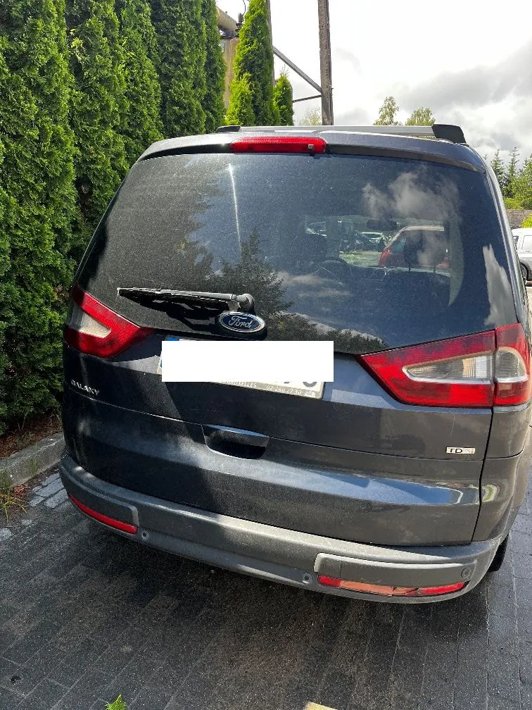 Tylny widok ciemnoszarego samochodu marki Ford Galaxy. Widoczna jest tylna szyba z wycieraczką, logo Ford na klapie bagażnika, tylne lampy oraz zderzak z odblaskami. Tablica rejestracyjna jest zakryta białym prostokątem. Po prawej stronie klapy znajduje się oznaczenie silnika TDCI. Samochód zaparkowany jest na kostce brukowej obok wysokiego, zielonego żywopłotu z tuj.