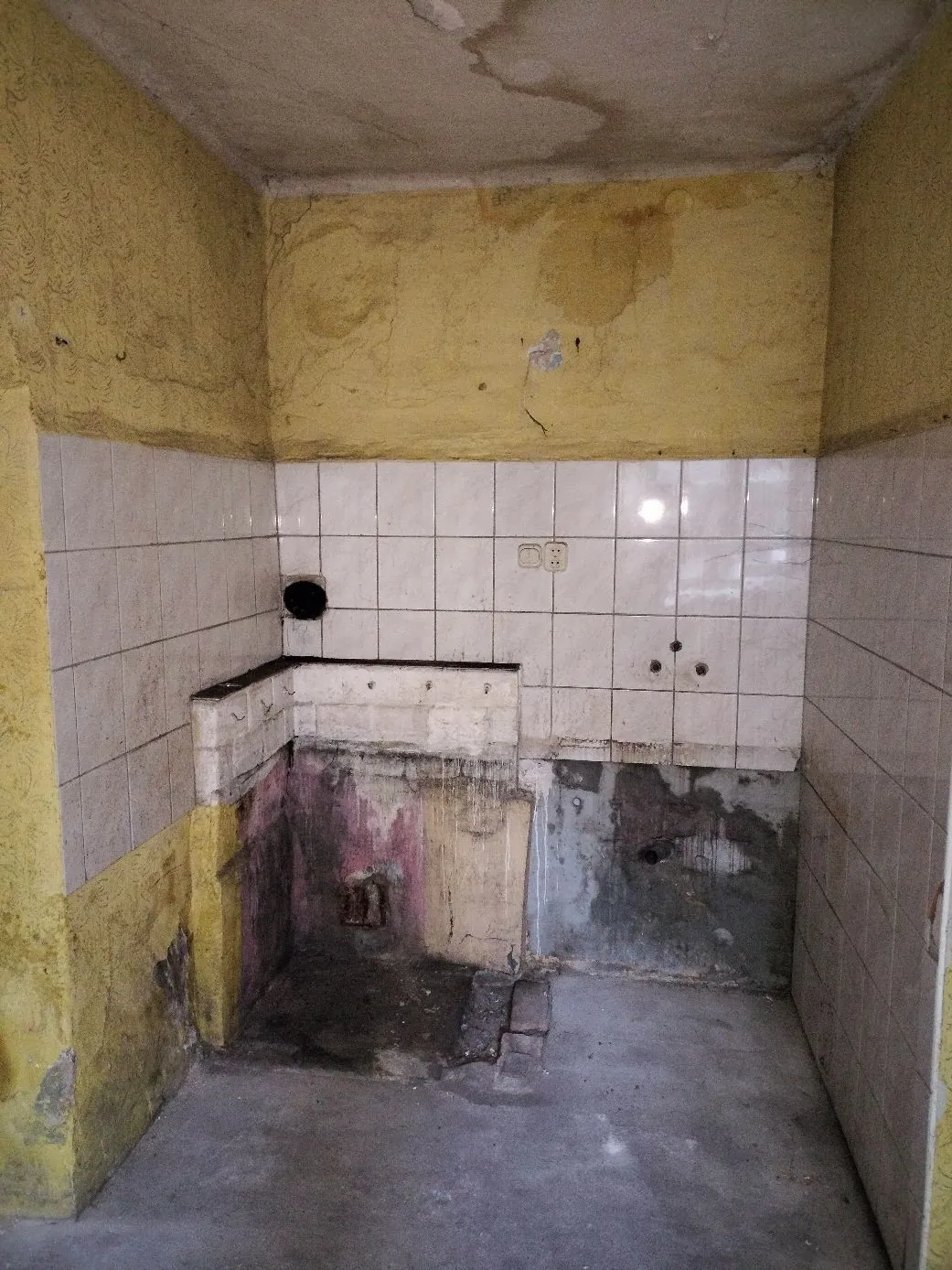 Wnętrze zniszczonego pomieszczenia, prawdopodobnie dawnej łazienki lub kuchni. Ściany są częściowo wyłożone białymi kafelkami, które są brudne i popękane. Powyżej kafelków ściany mają żółty, łuszczący się tynk. W centralnej części znajduje się betonowy cokół lub blat, pod którym widoczne są ślady wilgoci, pleśni i różowej farby. W ścianie z kafelkami znajduje się okrągły otwór wentylacyjny oraz dwa gniazdka elektryczne.