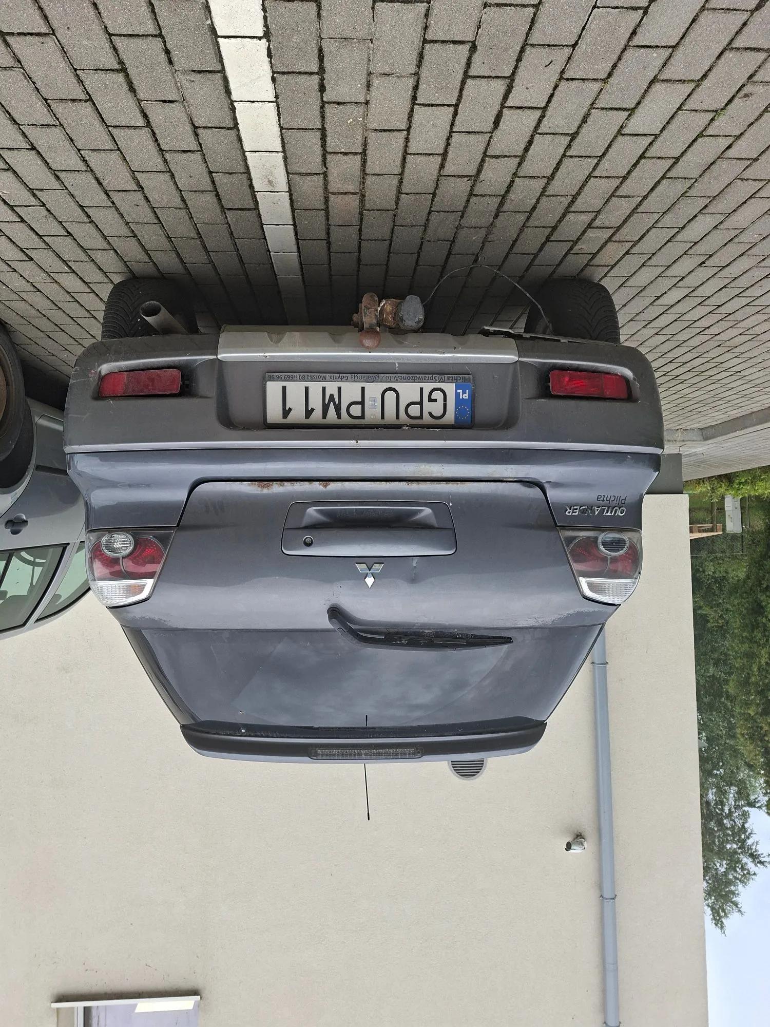Tylny widok szarego samochodu marki Mitsubishi Outlander, zaparkowanego na kostce brukowej. Na klapie bagażnika widoczne jest logo Mitsubishi oraz tablica rejestracyjna o numerze GPU PM11. Na lewym dole klapy znajduje się napis OUTLANDER i mniejszy napis pod nim. Obraz jest zorientowany poziomo, podczas gdy samochód i tekst na tablicy rejestracyjnej znajdują się w pionie.