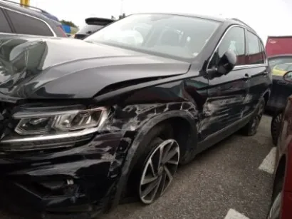 Czarny SUV z widocznymi uszkodzeniami na przedniej lewej stronie, w tym zgniecioną maską i zdeformowanym zderzakiem. Pojazd stoi na asfaltowym parkingu, obok innych samochodów, w tym czerwonego po prawej stronie. Tło zawiera fragmenty innych pojazdów i budynku z czerwoną ścianą. Brak czytelnego tekstu na obrazie.