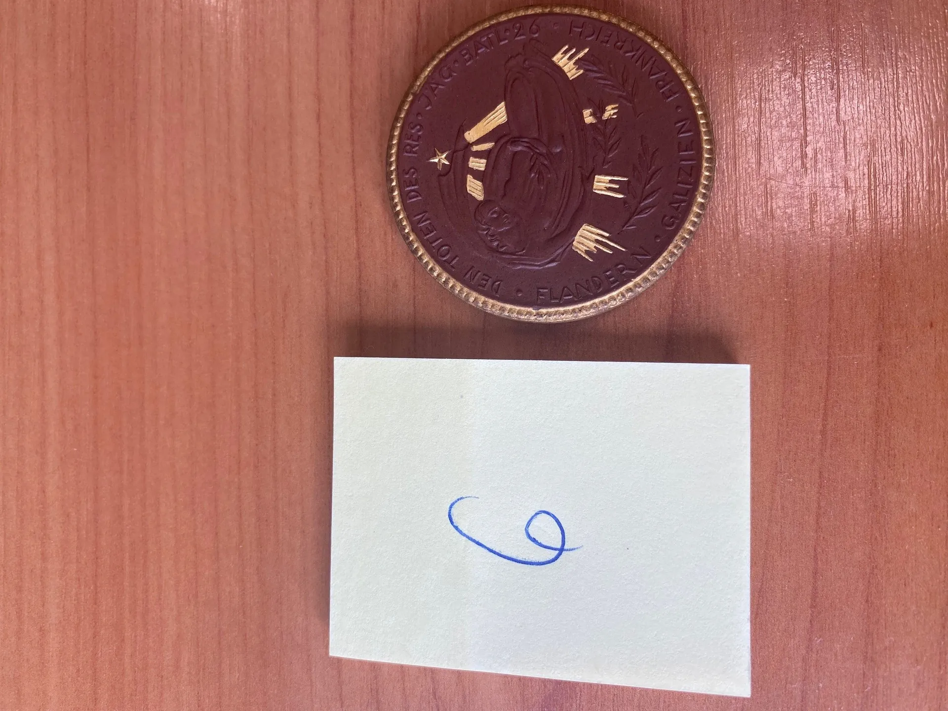 Biała kartka z niebieskim pismem ręcznym w kształcie pętli oraz ciemna moneta z złotymi inskrypcjami i postacią umieszczona na drewnianym tle.