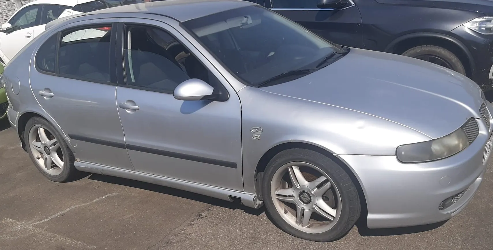 Srebrny samochód osobowy typu hatchback zaparkowany na asfaltowym placu. Pojazd jest widoczny z przodu i z boku, ma pięcioramienne felgi aluminiowe. W tle widoczne są fragmenty innych samochodów: białego po lewej stronie i czarnego po prawej.