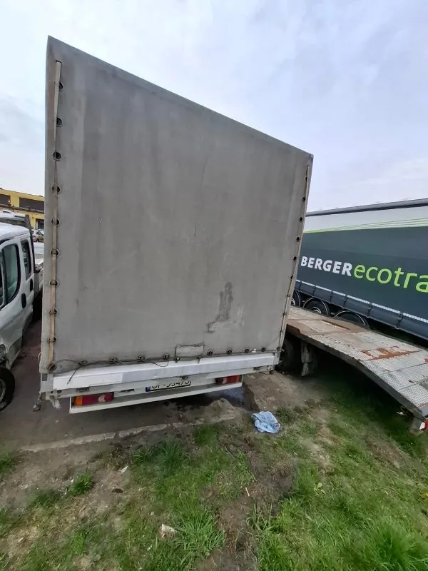 Zdjęcie przedstawia tył szarego naczepa ciężarowego z widocznymi czerwonymi światłami tylnymi i tablicą rejestracyjną. Po lewej stronie częściowo widoczny jest biały samochód dostawczy. Po prawej stronie znajduje się fragment innego pojazdu z napisem "BERGERecotra" na zielono-czarnym tle. Na ziemi, pokrytej trawą i błotem, leży niebieski przedmiot. Tło stanowi pochmurne niebo i fragment budynku w oddali.