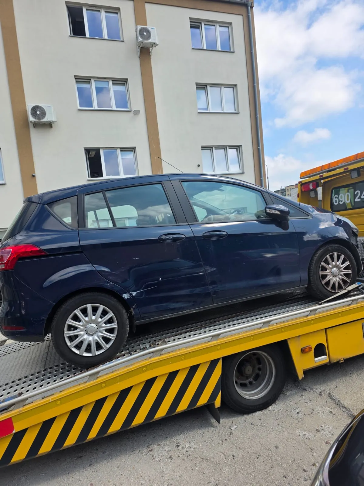 Ciemnoniebieski samochód osobowy typu hatchback zaparkowany na żółtej lawecie z czarno-żółtymi pasami ostrzegawczymi. W tle widoczny jest jasny, wielopiętrowy budynek mieszkalny z oknami i jednostkami klimatyzacji na ścianie zewnętrznej. Po prawej stronie widoczny jest fragment innego żółtego pojazdu z numerem 690 24.