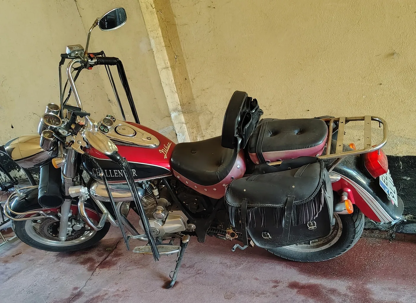 Czerwony motocykl typu cruiser zaparkowany bokiem na czerwonym podłożu przed żółtą ścianą. Motocykl posiada chromowane elementy, wysoki kierownicę, czarną skórzaną kanapę z oparciem dla kierowcy oraz czarną torbę sakwę z frędzlami zamocowaną z tyłu. Na baku widnieje napis 'Challenger'.