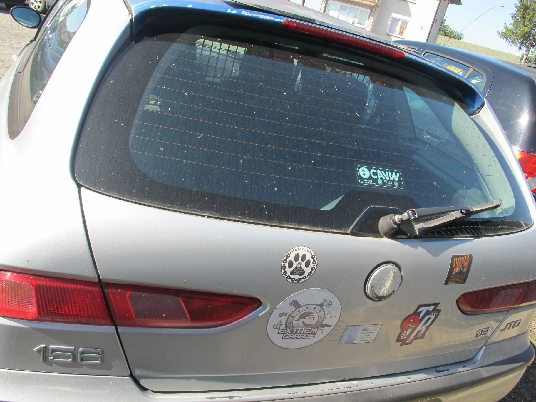 Tylna część srebrnego samochodu marki Alfa Romeo, model 156. Widoczna jest brudna tylna szyba z wycieraczką oraz naklejką 'CAIW'. Na klapie bagażnika znajdują się liczne naklejki, w tym okrągła z napisem 'MotoLaps' i łapą zwierzęcia oraz większa z napisem 'EXTREME GARAGE'. W lewym dolnym rogu widnieje oznaczenie modelu '156', a po prawej stronie napis 'JTD'.