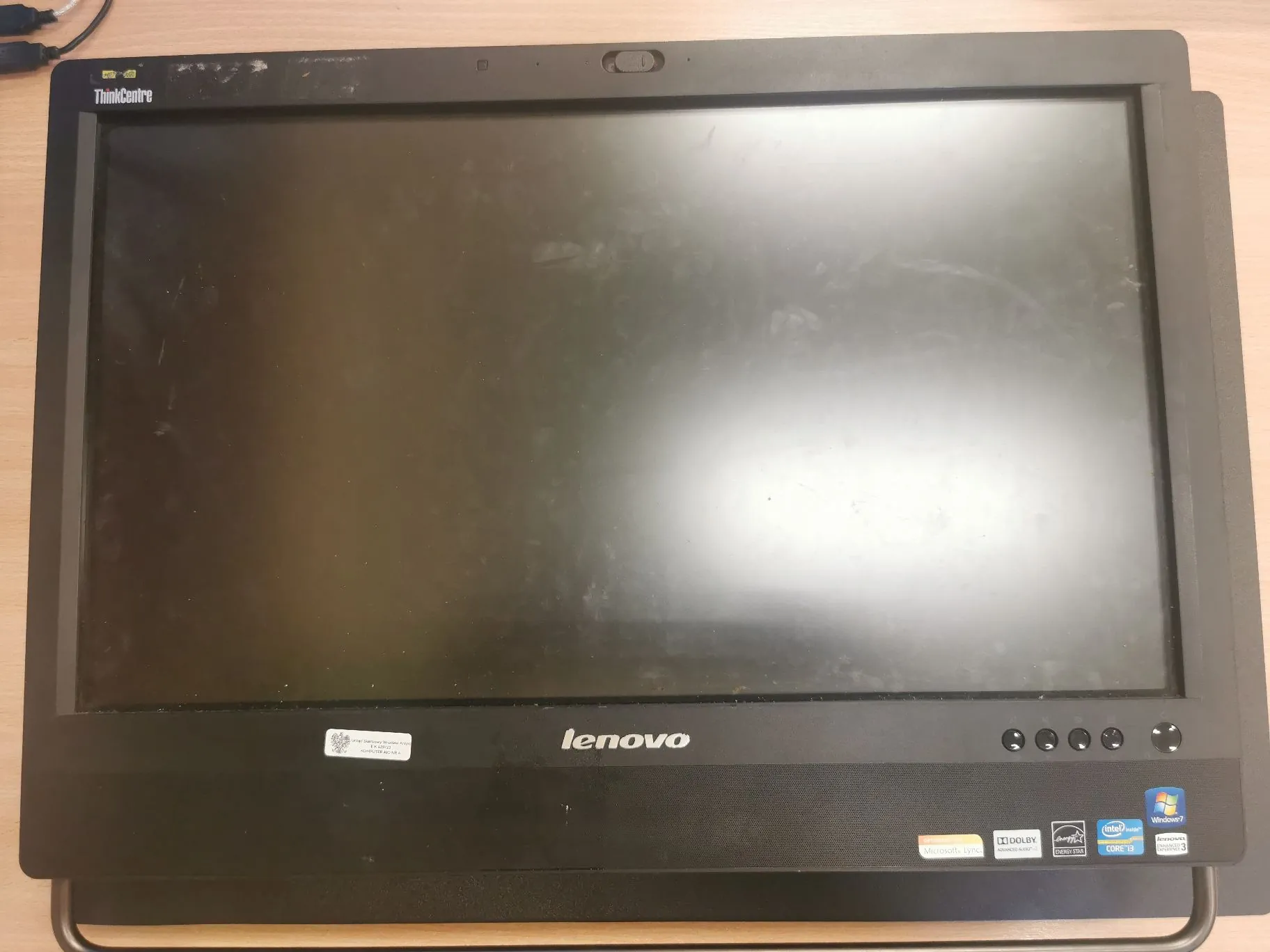 Czarny monitor marki Lenovo ThinkCentre z wyłączonym ekranem. W lewym górnym rogu znajduje się napis 'ThinkCentre', a na dole na ramce logo 'lenovo'. W prawym dolnym rogu widoczne są przyciski sterujące oraz naklejki z logo Intel Core i3, Windows 7 oraz Dolby. W lewym dolnym rogu znajduje się mała biała etykieta z kodem kreskowym.
