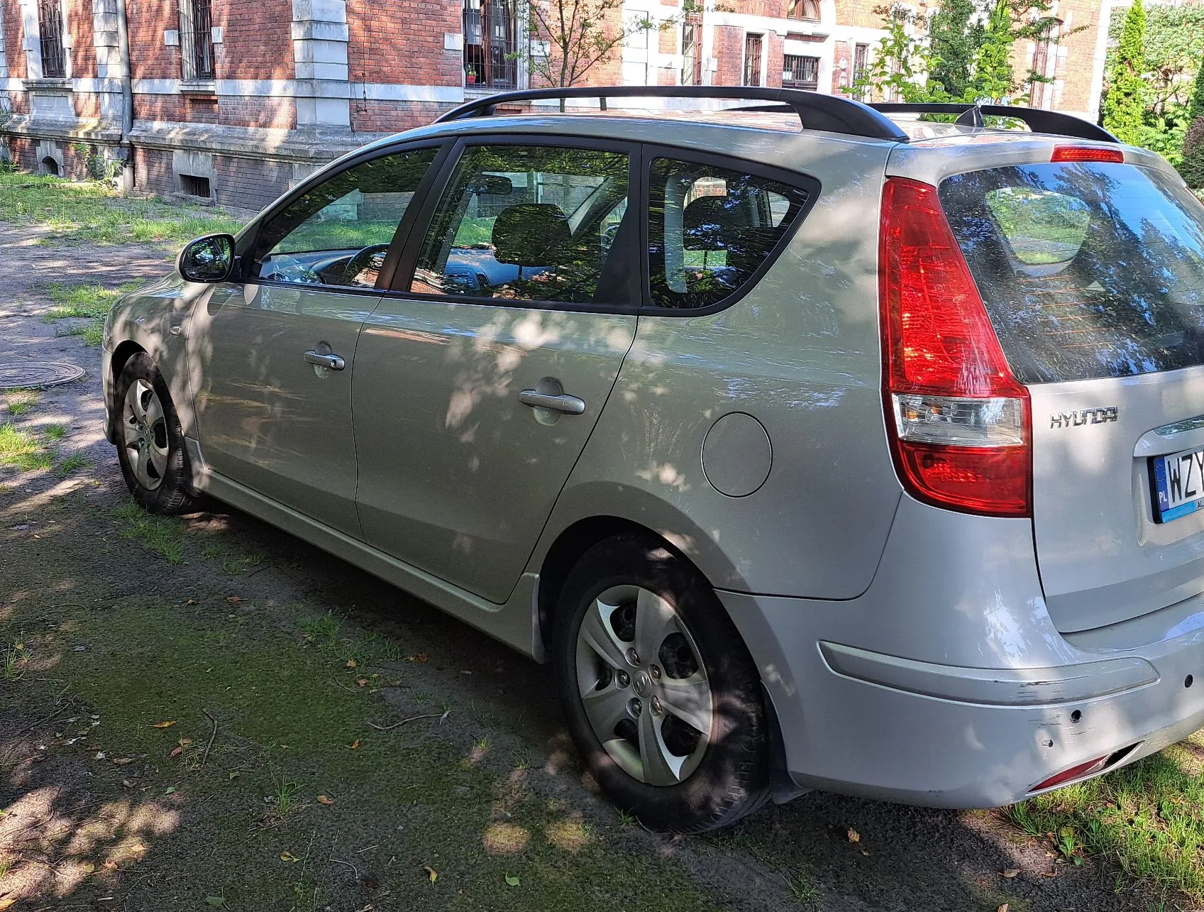 Srebrny samochód marki Hyundai w nadwoziu kombi, zaparkowany na trawiasto-piaszczystym podłożu przed ceglanym budynkiem. Widoczny jest tył i prawy bok pojazdu, relingi dachowe oraz fragment tablicy rejestracyjnej zaczynający się od liter WZ.
