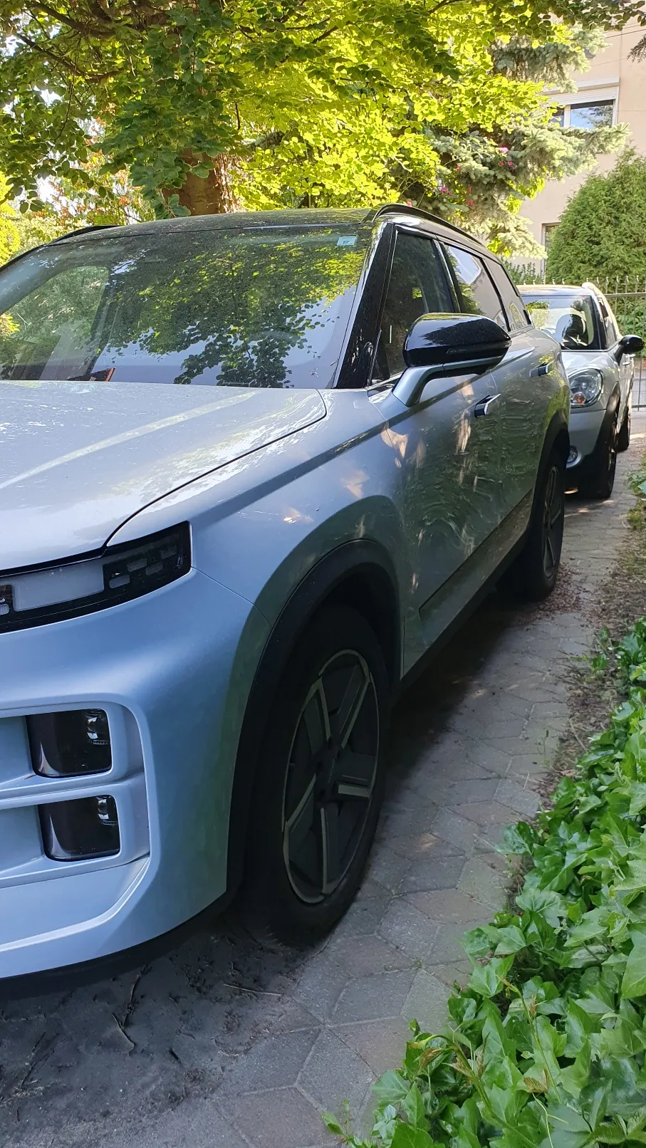 Srebrny samochód typu SUV zaparkowany na kostce brukowej obok zielonego żywopłotu. Nad pojazdem znajdują się rozłożyste drzewa z zielonymi liśćmi. W tle widoczny jest fragment innego, białego samochodu oraz ściana budynku.