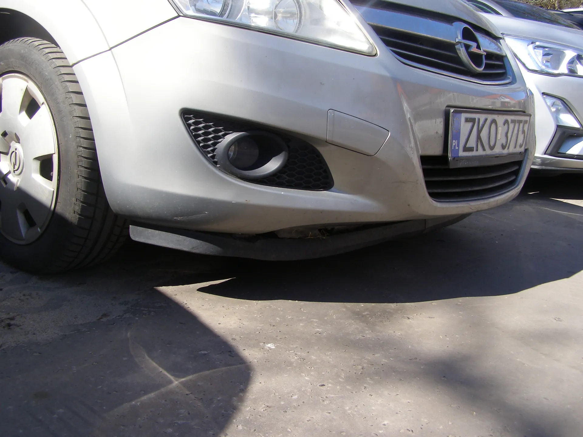 Przód srebrnego samochodu z widoczną tablicą rejestracyjną PL ZKO 3715, atrapą chłodnicy z logo, reflektorem i częścią koła. W tle fragment innego srebrnego samochodu. Samochody zaparkowane na betonowej powierzchni, widoczne cienie.