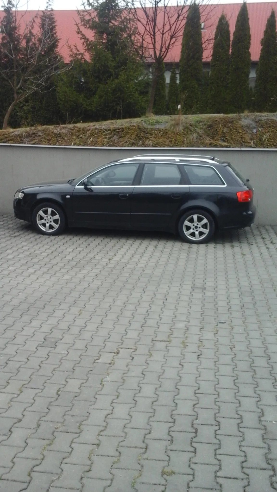 Czarny samochód typu kombi zaparkowany na powierzchni z betonowych kostek. Po lewej stronie samochodu znajduje się pas trawy z drzewami i czerwony budynek na tle. Samochód ma srebrne felgi i widoczne czerwone światło tylnego.