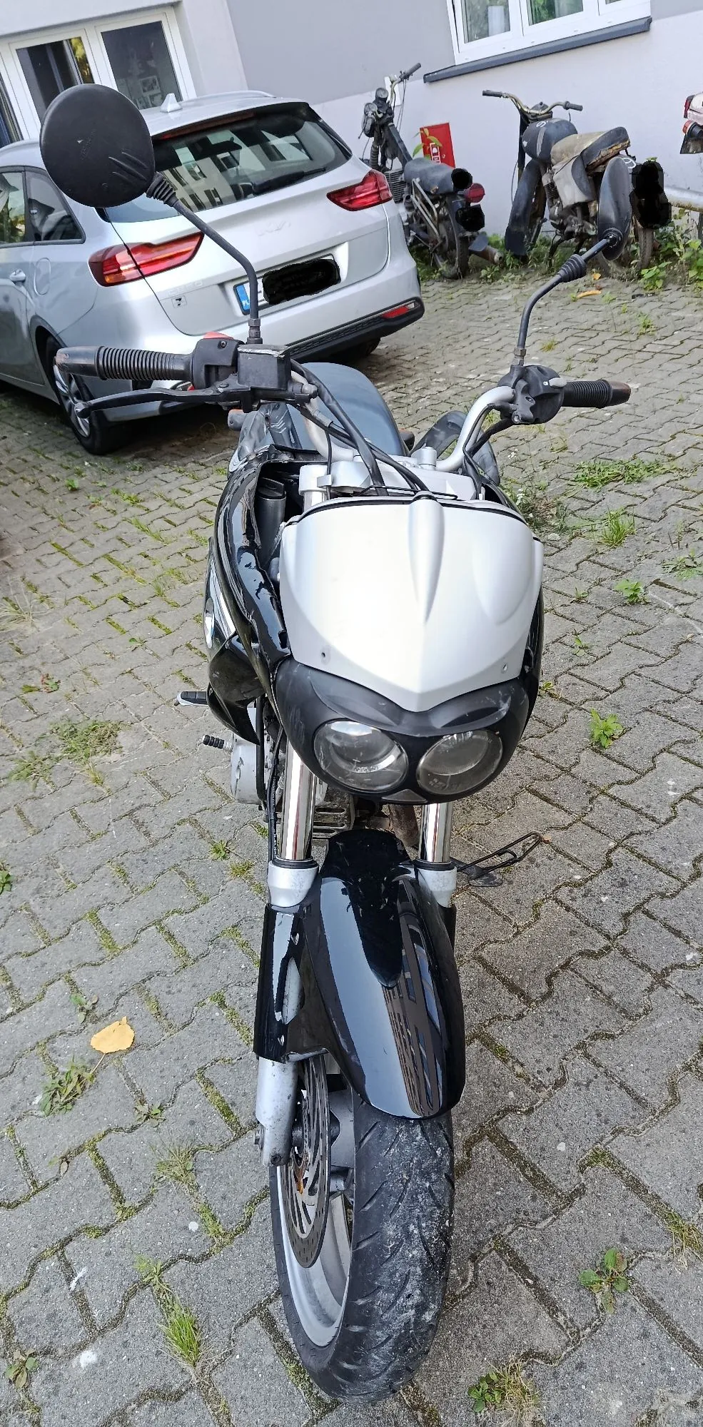Widok z przodu na czarno-srebrny motocykl zaparkowany na kostce brukowej. Motocykl ma srebrną owiewkę nad podwójnym reflektorem oraz czarny błotnik przedni. W tle widoczny jest srebrny samochód oraz inny motocykl zaparkowany przy ścianie budynku.