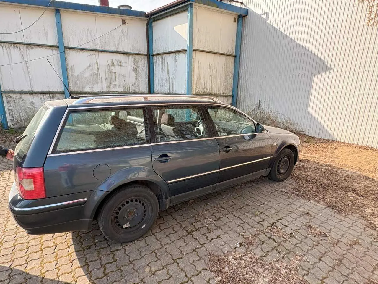 Ciemnoszary samochód typu kombi zaparkowany na brukowanej powierzchni. Pojazd ma dachowy bagażnik, czarne felgi i widoczne wnętrze z siedzeniami. W tle budynek przemysłowy z wyblakłymi, zniszczonymi panelami i niebieskimi elementami konstrukcyjnymi. Na ziemi sucha trawa i liście. Brak czytelnego tekstu.