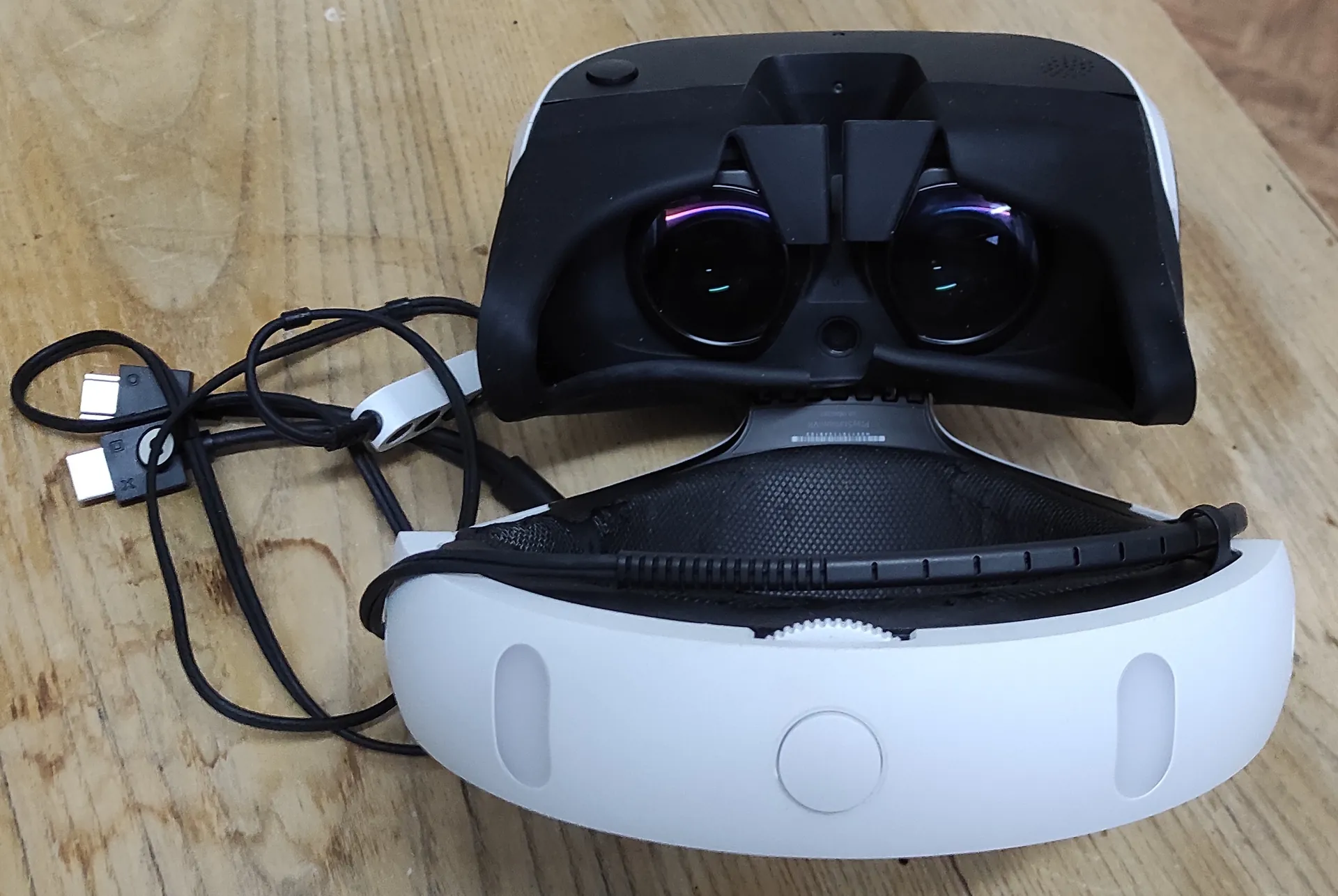 Gogle wirtualnej rzeczywistości (VR) w kolorach białym i czarnym, leżące na drewnianym blacie. Urządzenie jest rozłożone, ukazując soczewki oraz biały pasek mocujący. Obok znajduje się czarny kabel z dwoma wtyczkami USB.