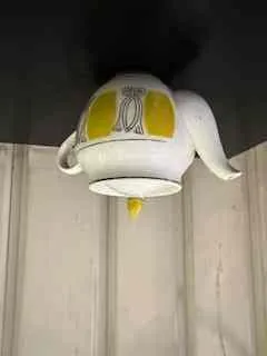 Biały czajnik z żółtymi i szarymi wzorami, z żółtym uchwytem na pokrywce, umieszczony na ciemnej powierzchni. W tle widoczne są jasne, poziome półki lub schody.