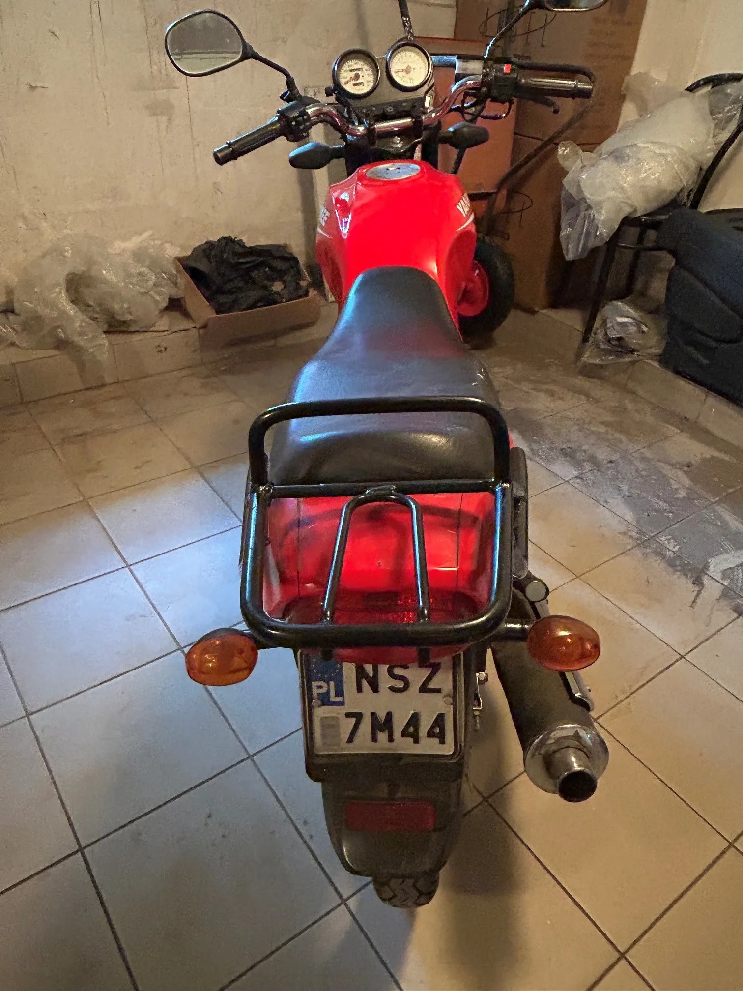 Czerwony motocykl zaparkowany w pomieszczeniu z kafelkami na podłodze. Widok z tyłu pojazdu, ukazujący czarne siedzenie, metalowy bagażnik, tylne światło oraz tablicę rejestracyjną o numerze NSZ 7M44 z oznaczeniem PL. Po prawej stronie widoczna jest rura wydechowa, a po bokach dwa pomarańczowe kierunkowskazy. W tle znajdują się kartony i przedmioty przykryte folią.