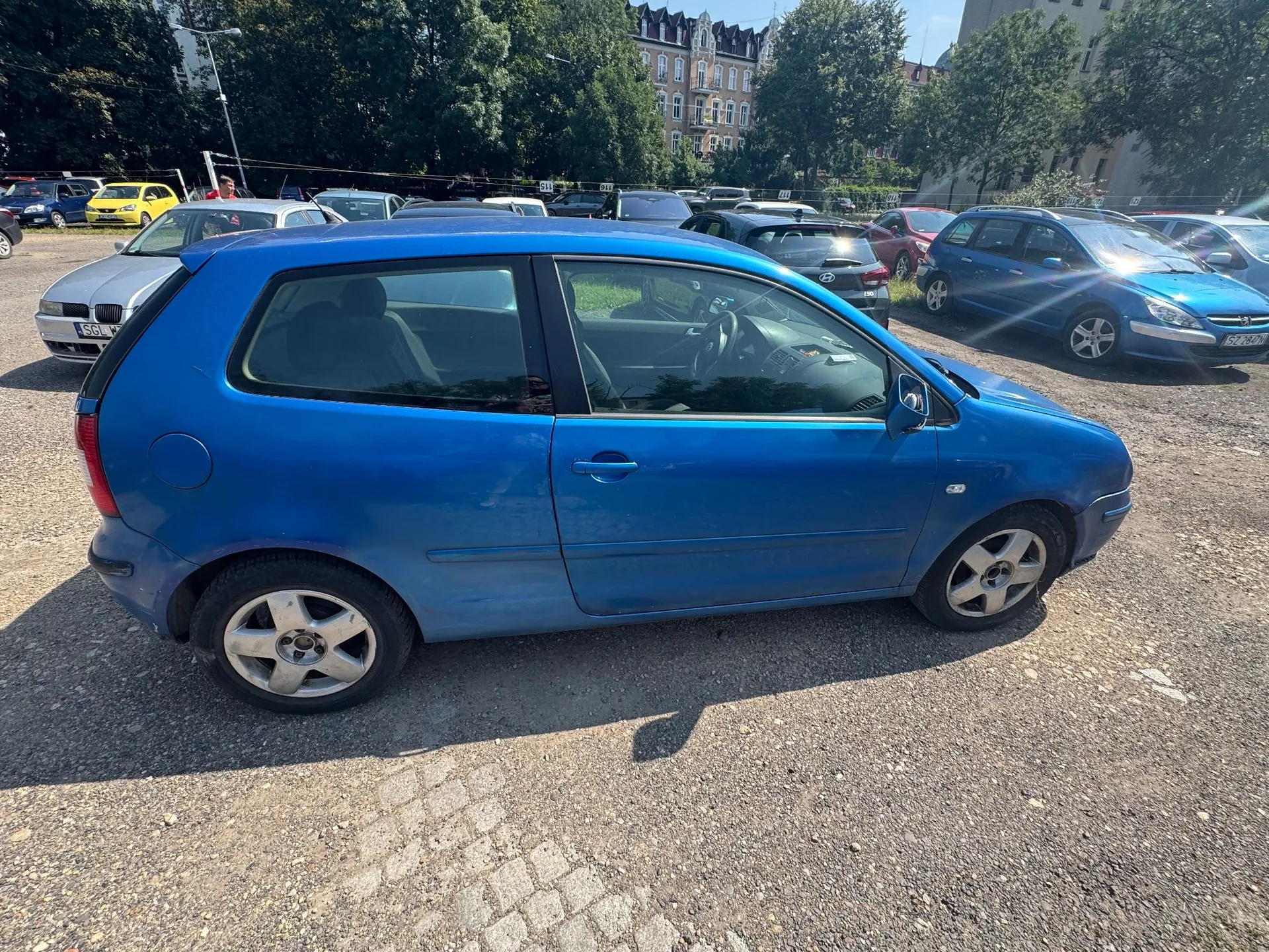 Niebieski, trzydrzwiowy samochód osobowy zaparkowany bokiem na żwirowym parkingu. W tle widoczne są inne zaparkowane samochody w różnych kolorach, zielone drzewa oraz fragmenty budynków mieszkalnych.