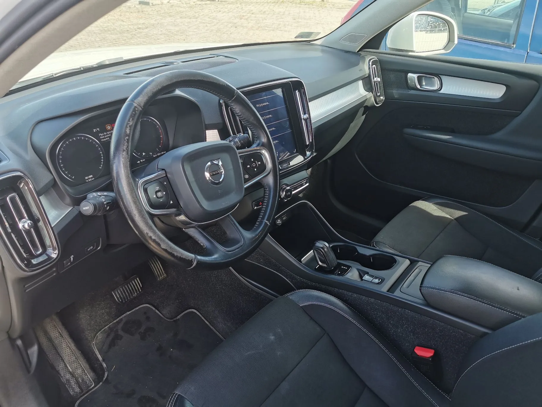 Wnętrze samochodu marki Volvo z perspektywy miejsca kierowcy. Widoczna jest czarna skórzana kierownica z logo Volvo w centrum, deska rozdzielcza z cyfrowymi zegarami, centralny ekran dotykowy oraz konsola środkowa z lewarkiem zmiany biegów i podłokietnikiem. Fotele są wykonane z ciemnego materiału, a na podłodze znajdują się czarne dywaniki.