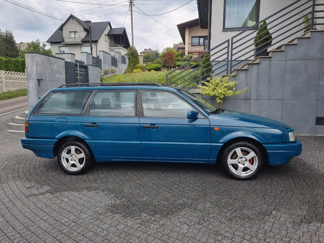 Niebieski samochód typu kombi zaparkowany na brukowanej powierzchni. W tle widoczne są domy mieszkalne z dachami, betonowa ściana z metalowymi schodami, zieleń oraz chmurne niebo. Samochód ma srebrne felgi i dachowy bagażnik. Wokół znajdują się ogrodzenia, krzewy i drzewa.