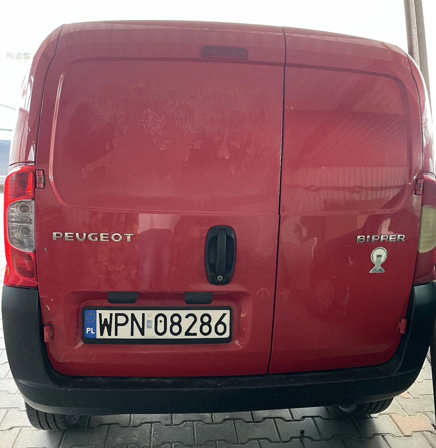 Tylny widok czerwonego samochodu marki Peugeot Bipper. Na klapie bagażnika widoczne są napisy 'PEUGEOT' po lewej stronie oraz 'BIPPER' po prawej stronie wraz z małym logo. Samochód posiada polską tablicę rejestracyjną o numerze WPN 08286. Karoseria w górnej części jest zmatowiała i posiada ślady zużycia lakieru.