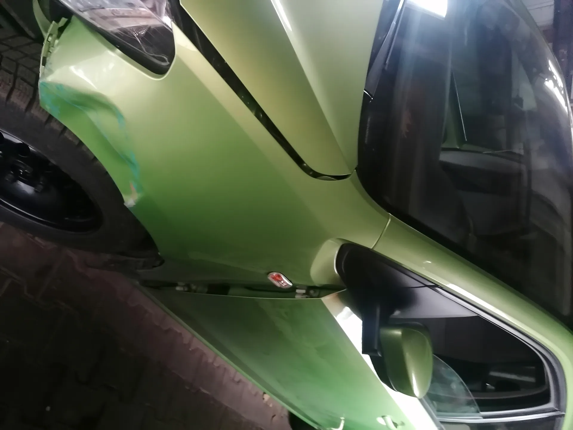 Zielony samochód z widocznym uszkodzeniem na przednim zderzaku, gdzie odsłonięta jest niebieska farba. Drzwi pasażerskie są otwarte, a lusterko boczne widoczne. Samochód zaparkowany na brukowanej powierzchni w pomieszczeniu zadaszonym. Szyby przyciemnione, widoczne reflektory i czarne felgi.