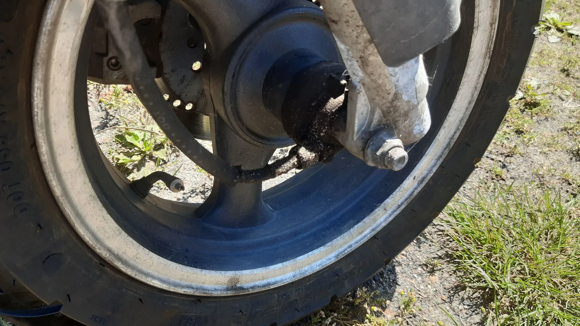 Zbliżenie na koło motocyklowe z widocznym układem hamulcowym, tarczą oraz wahaczem. Koło jest zamontowane w ramie, a w tle widoczna jest trawa i ziemia.