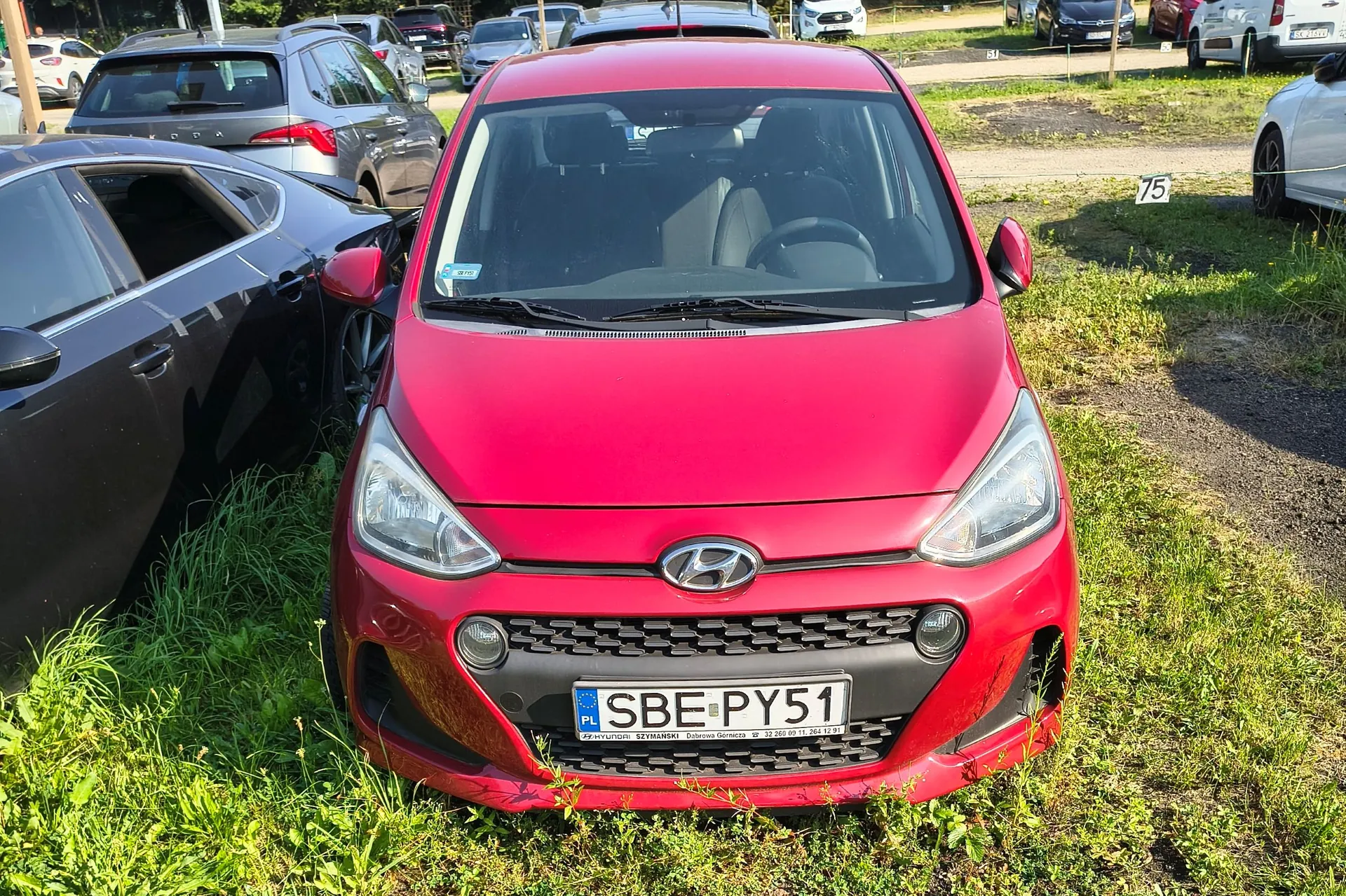 Czerwony samochód marki Hyundai zaparkowany na trawiastym terenie. Widoczna jest przednia część pojazdu z tablicą rejestracyjną o numerze SBE PY51. Po lewej stronie znajduje się czarny samochód z uszkodzeniami, a w tle inne zaparkowane auta.