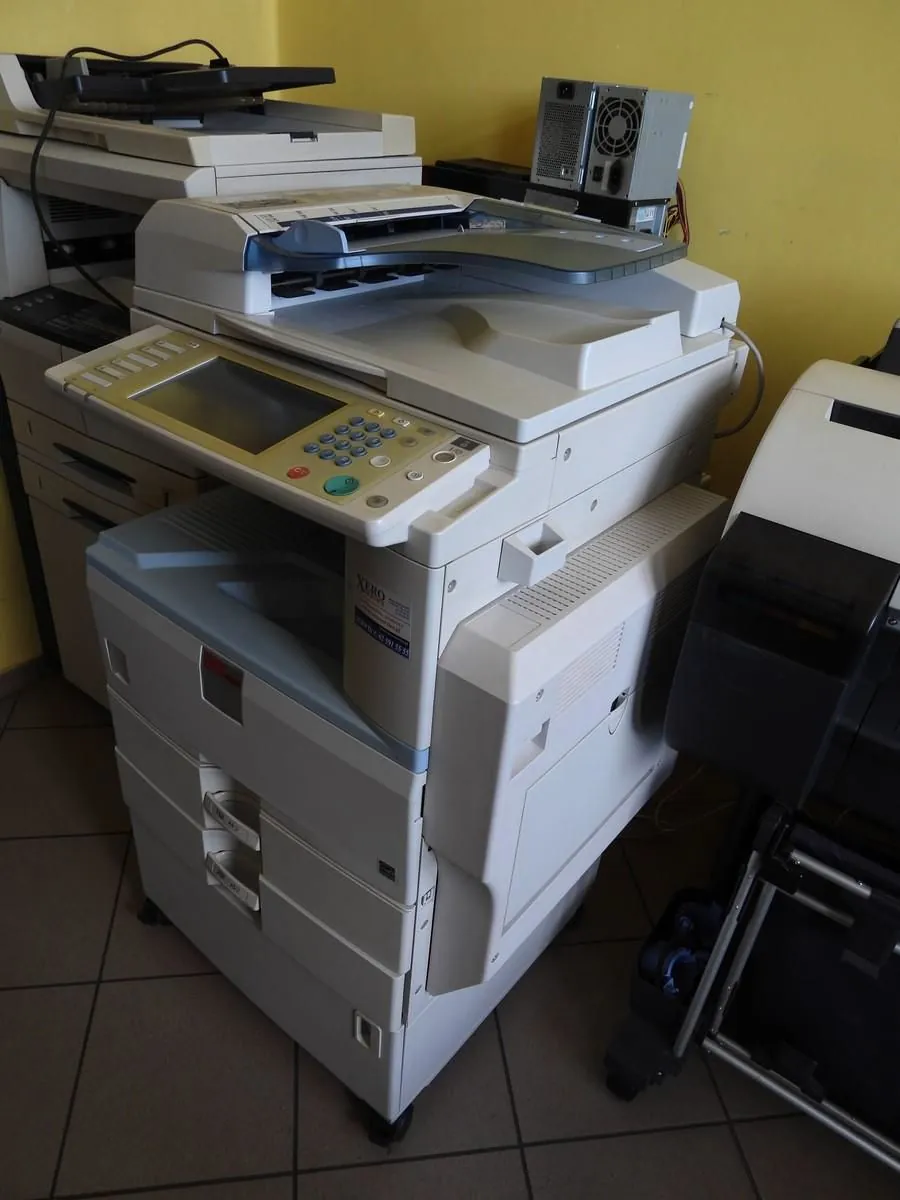 Duża, biała wielofunkcyjna drukarka biurowa marki Xerox z panelem sterowania, ekranem i wieloma przyciskami. Urządzenie stoi na płytkach ceramicznych w pomieszczeniu z żółtą ścianą. W tle widoczne są inne urządzenia biurowe oraz zasilacz komputerowy leżący na górnej powierzchni.