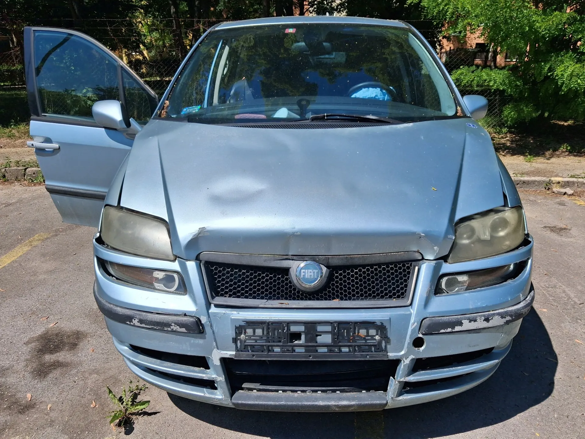 Srebrny samochód marki Fiat widziany z przodu. Auto jest uszkodzone: maska jest wgnieciona, a przedni zderzak posiada liczne zarysowania, odpryski lakieru oraz wgniecenia. Lewe przednie drzwi są otwarte. Reflektory są zmatowiałe. Na środku grilla znajduje się okrągłe logo FIAT.