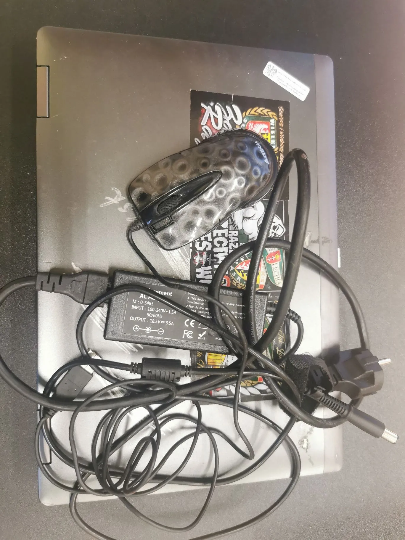 Zdjęcie z góry przedstawiające zamknięty, ciemnoszary laptop z naklejkami na obudowie. Na laptopie leży czarna mysz komputerowa z wzorem w kropki oraz zasilacz z kablem, który jest częściowo splątany. Na zasilaczu widoczna jest etykieta z parametrami technicznymi: 'INPUT: 100-240V~1.5A 50/60Hz' oraz 'OUTPUT: 18.5V == 3.5A'.
