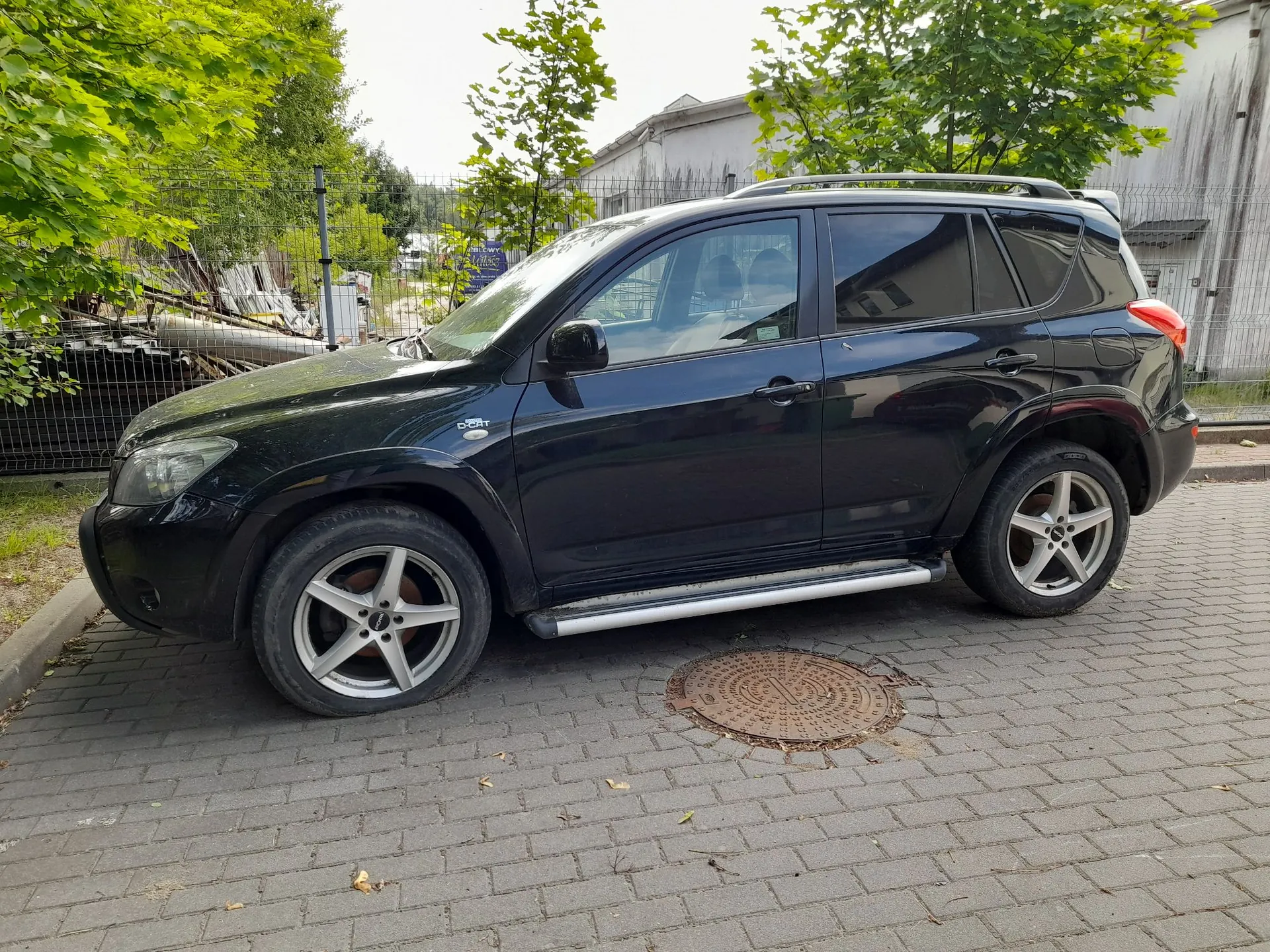 Czarny samochód typu SUV zaparkowany bokiem na kostce brukowej. Pojazd posiada srebrne felgi aluminiowe z pięcioma ramionami oraz srebrne stopnie boczne. W tle widoczne są zielone drzewa, ogrodzenie z siatki oraz fragmenty budynków przemysłowych. Na ziemi, obok samochodu, znajduje się okrągły właz kanalizacyjny.
