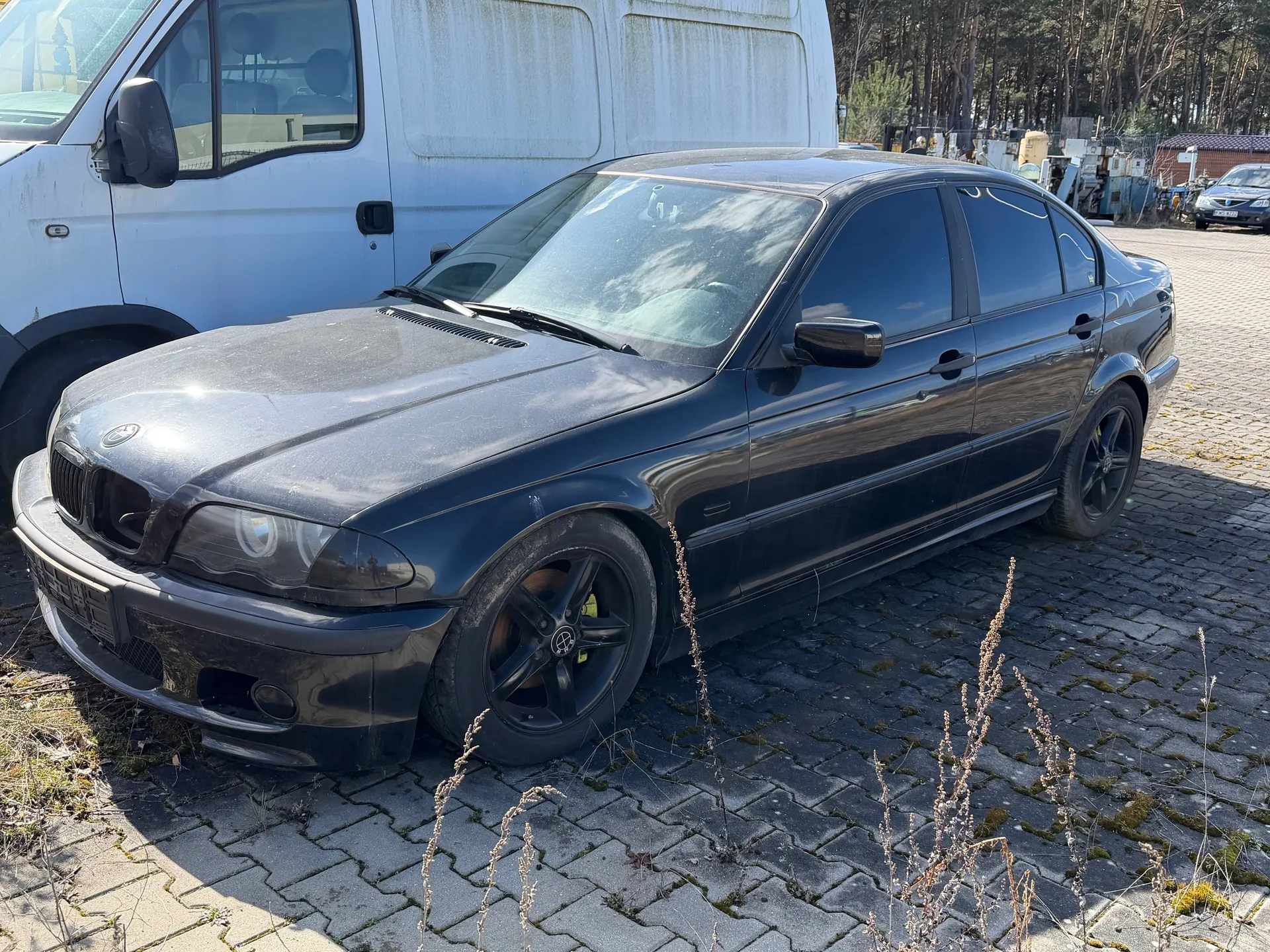 Czarny samochód osobowy marki BMW, model sedan, zaparkowany na brukowanej powierzchni. Po lewej stronie widoczny biały furgon. W tle drzewa, inne pojazdy oraz budynki. Samochód ma czarne felgi, przednią maskę z charakterystyczną atrapą chłodnicy i reflektory. Na przednim zderzaku widoczna tablica rejestracyjna. Powierzchnia brukowa pokryta jest suchą trawą i mchem. Oświetlenie słoneczne, cienie wskazują na jasny dzień.