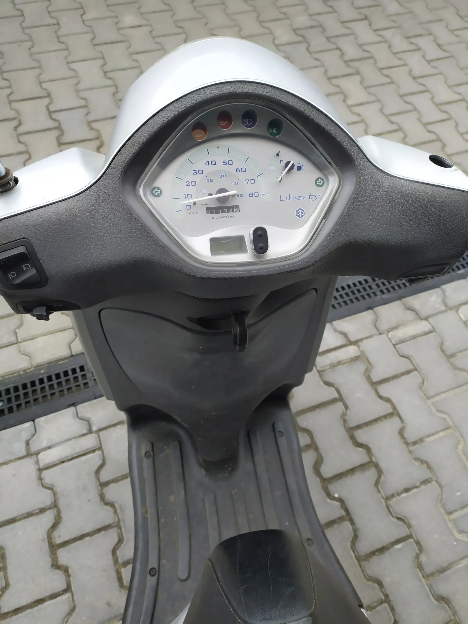 Widok z perspektywy kierowcy na kokpit srebrnego skutera zaparkowanego na kostce brukowej. Widoczny jest licznik prędkości z napisem 'Liberty', wskaźnik paliwa oraz czarna obudowa kierownicy i podnóżek.