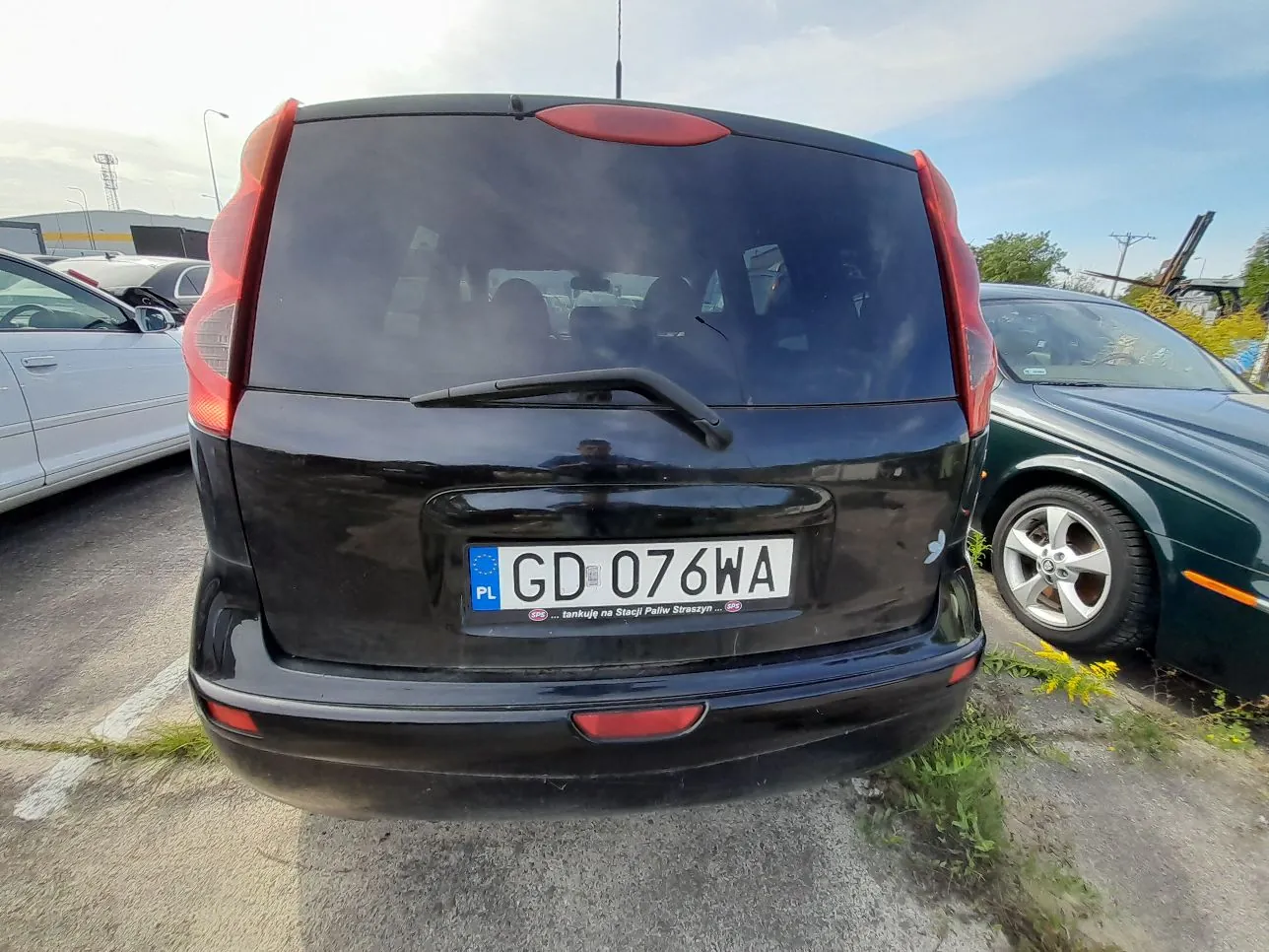 Tylny widok czarnego samochodu typu minivan zaparkowanego na placu. Pojazd posiada polską tablicę rejestracyjną o numerze GD 076WA. Pod numerem rejestracyjnym znajduje się ramka z napisem „... tankuj na Stacji Paliw Streszyn ...”. Po lewej stronie widoczny jest fragment srebrnego samochodu, a po prawej ciemnozielonego samochodu.