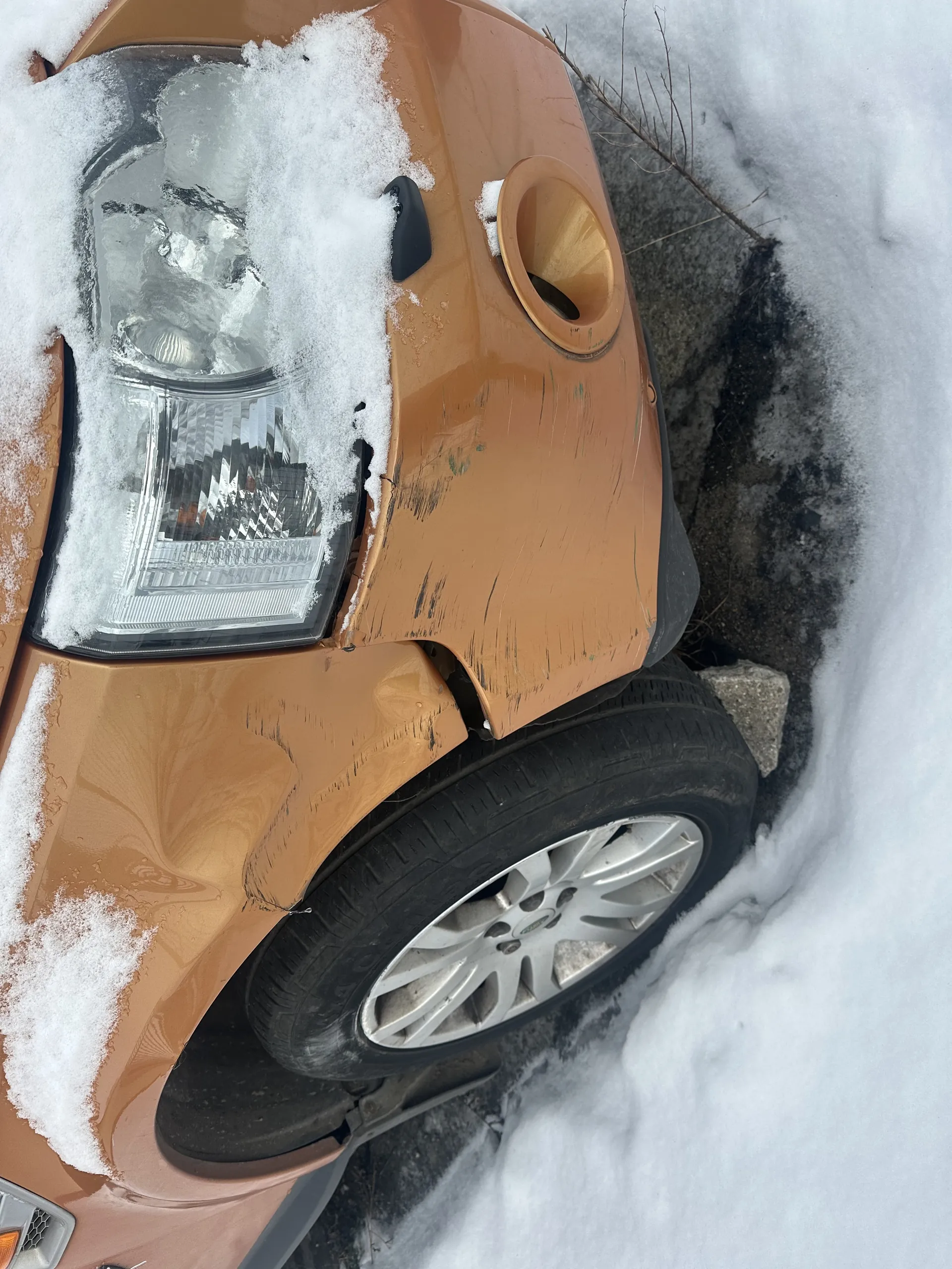 Pomarańczowy samochód z przodu, z widocznym reflektorem i zniszczonym zderzakiem. Na masce i reflektorze leży śnieg. Opona z srebrną felgą, na zderzaku widoczne zadrapania i uszkodzenia. Śnieg na ziemi, częściowo odsłonięta nawierzchnia drogi.