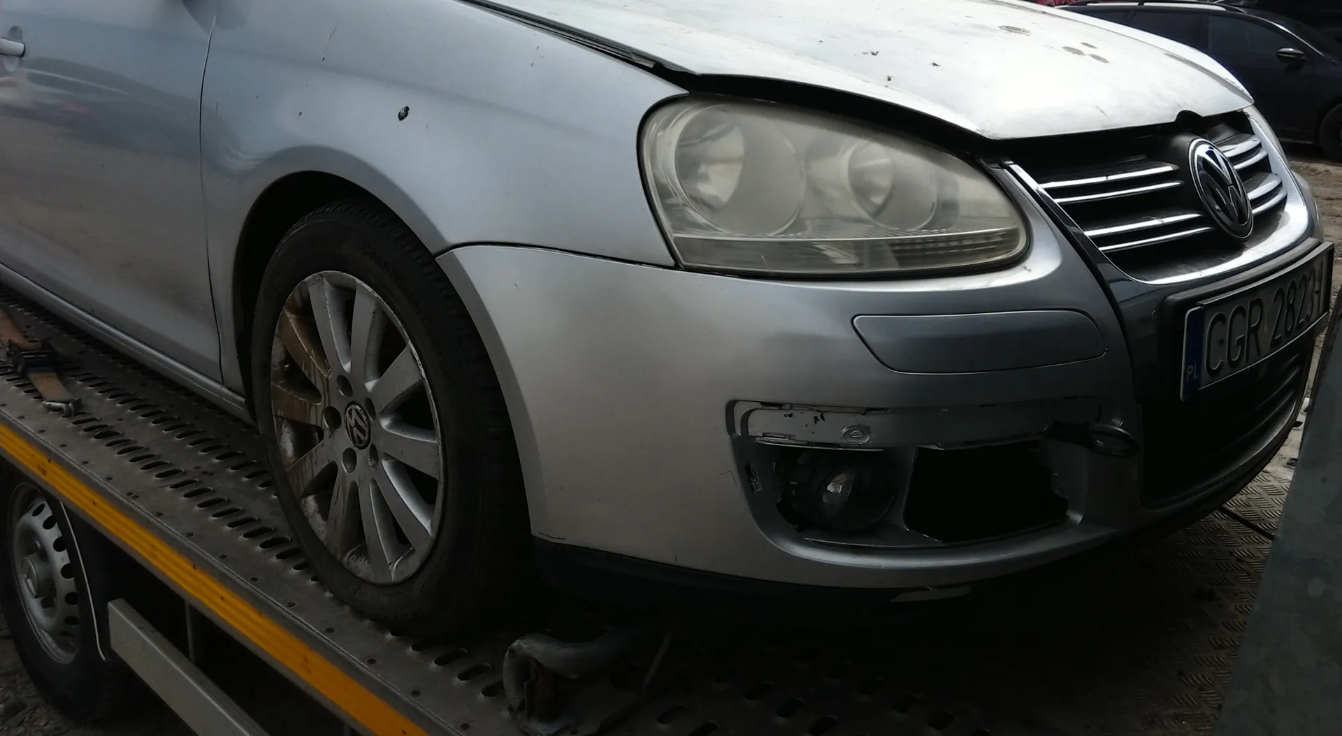 Srebrny samochód marki Volkswagen na platformie holownika. Widoczna przód pojazdu z uszkodzonym zderzakiem, reflektorem, atrapą chłodnicy z logo VW oraz tablicą rejestracyjną CGR 2823 z oznaczeniem PL. Koło przednie z felgą i oponą marki Continental. Platforma holownika z żółtą linią i perforowaną powierzchnią. W tle częściowo widoczny inny pojazd.