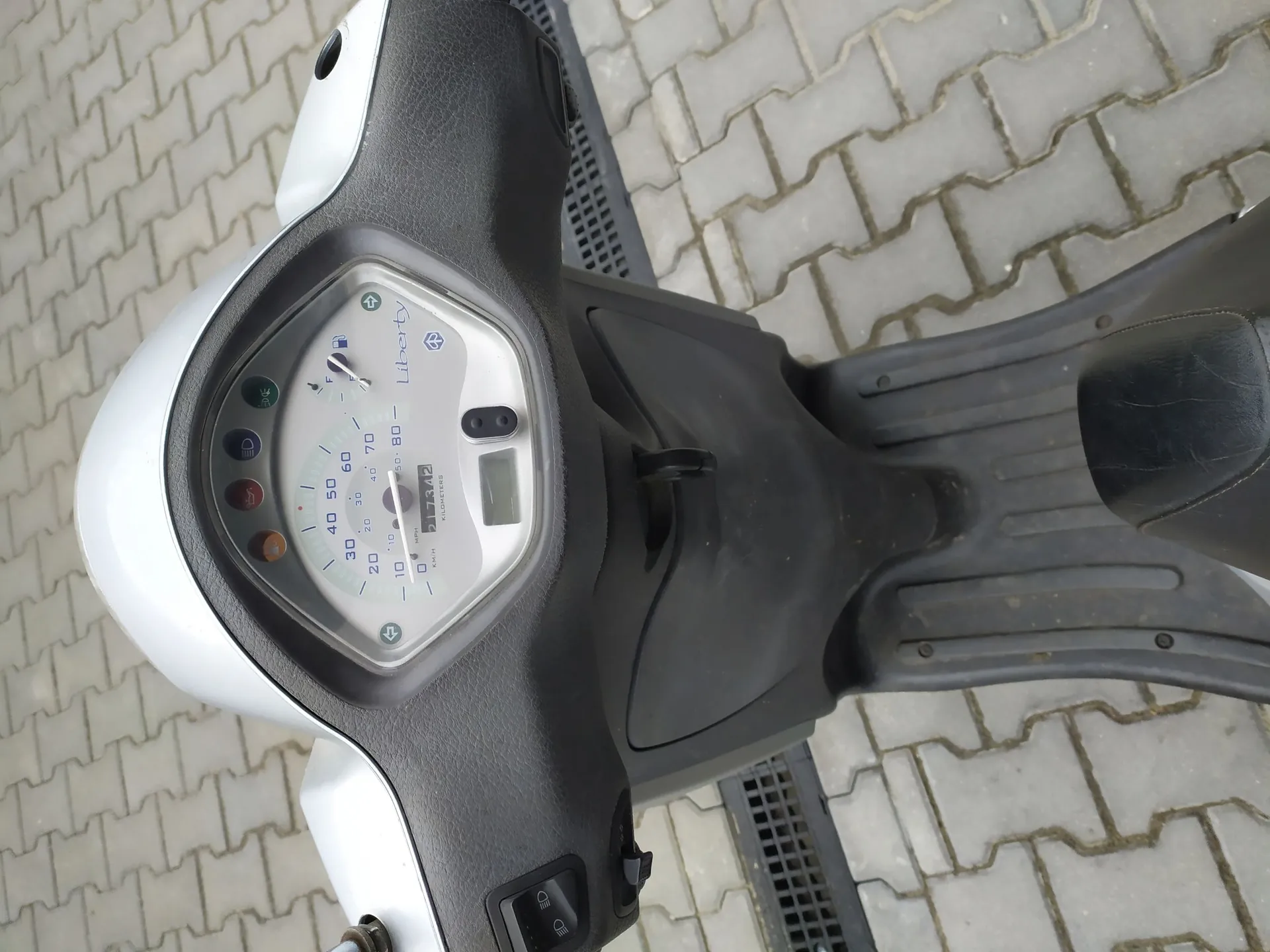 Widok z perspektywy kierowcy na kokpit i przód srebrnego skutera marki Liberty. Widoczny jest licznik prędkości z oznaczeniami od 0 do 80 km/h, wskaźnik paliwa oraz kontrolki. Skuter stoi na szarym chodniku z kostki brukowej.