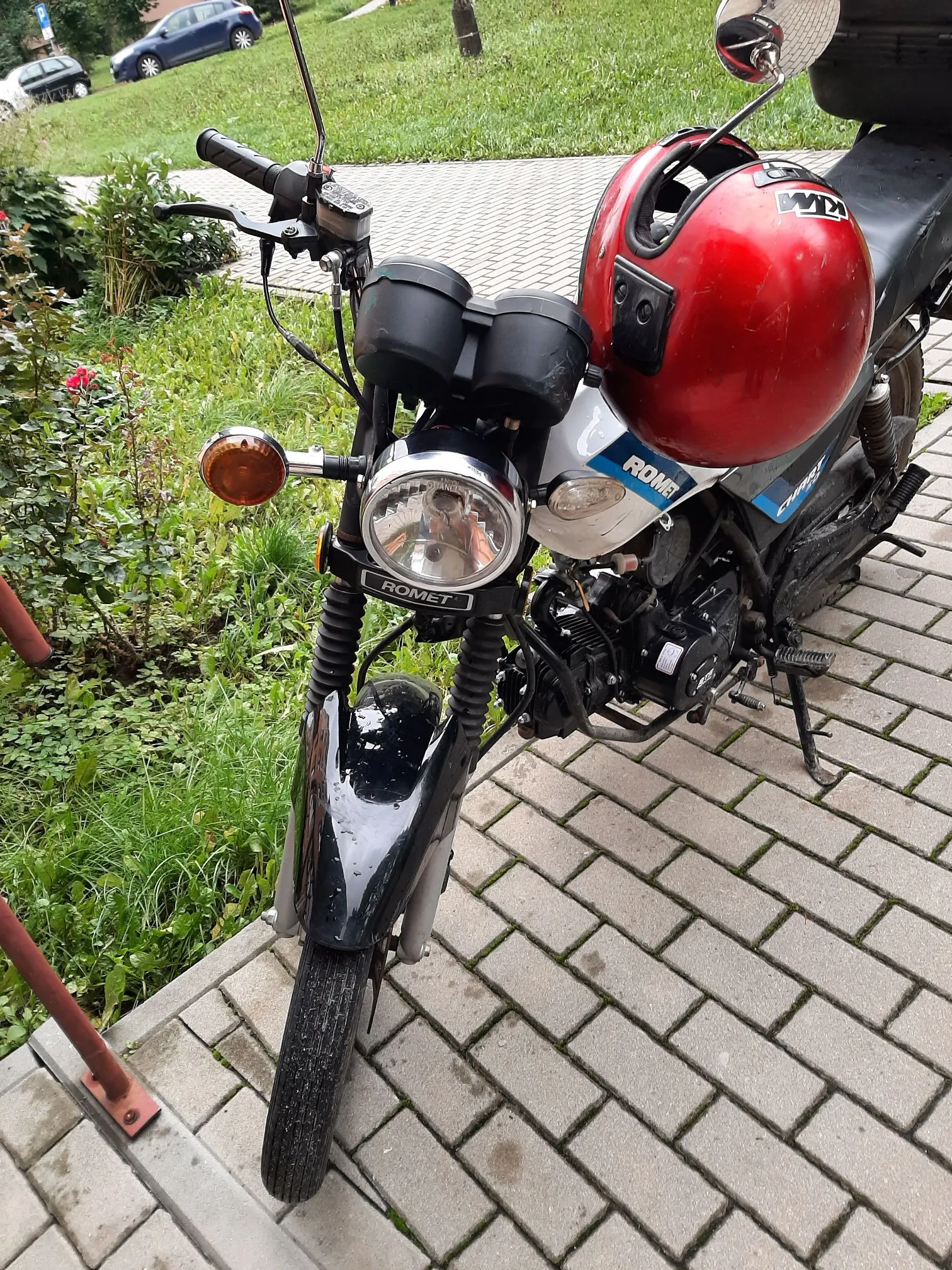 Zbliżenie na przód czarnego motocykla marki Romet, zaparkowanego na kostce brukowej. Na siedzeniu motocykla spoczywa czerwony kask. Widoczny jest okrągły reflektor z napisem ROMET, kierunkowskazy oraz przednie zawieszenie. W tle znajduje się zielona trawa i fragmenty samochodów.