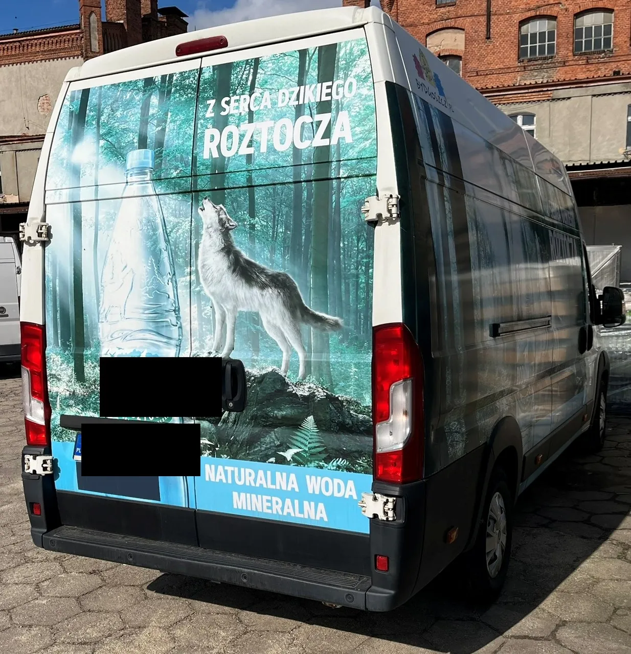 Tył i bok białego samochodu dostawczego oklejonego reklamą wody mineralnej. Na grafice widoczny jest las, butelka wody oraz wilk stojący na skale i patrzący w górę. Na górze znajduje się napis: 'Z SERCA DZIKIEGO ROZTOCZA', a na dole: 'NATURALNA WODA MINERALNA'. Część tylnych drzwi jest zakryta czarnymi polami. Samochód zaparkowany jest na kostce brukowej przed ceglanym budynkiem.