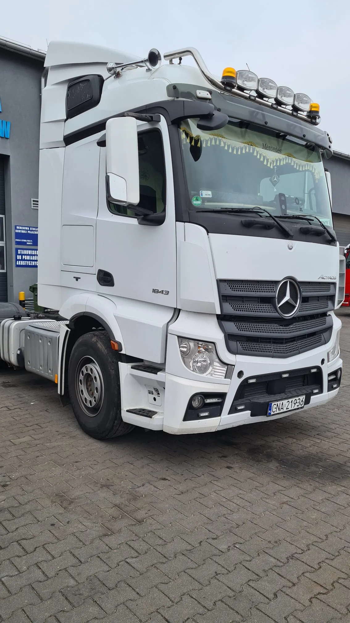 Biała ciężarówka marki Mercedes-Benz Actros zaparkowana na kostce brukowej. Pojazd posiada dodatkowe oświetlenie na dachu, tablicę rejestracyjną GNA 21936 oraz oznaczenie modelu 1843 na boku kabiny. W tle widoczny jest szary budynek z niebieską tablicą informacyjną z tekstem: 'STANOWISKO DO POMIARU AERODYNAMICZNEGO'.
