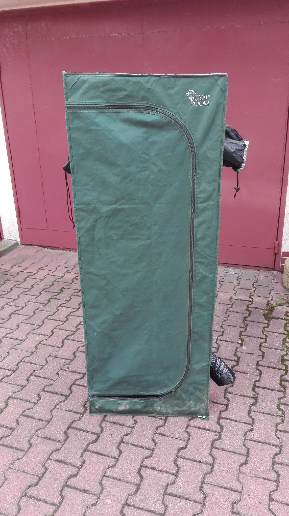 Zielony, materiałowy pokrowiec transportowy stojący pionowo na kostce brukowej na tle bordowej ściany. W prawym górnym rogu pokrowca znajduje się biały napis „ROYAL ROOM”. Pokrowiec ma zamek błyskawiczny biegnący wzdłuż krawędzi.